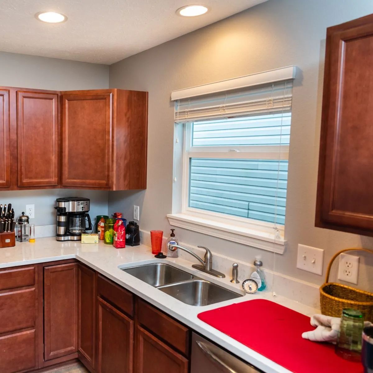 Kitchen with wooden cabinets, double sink, window, coffee maker, and a red drying mat.