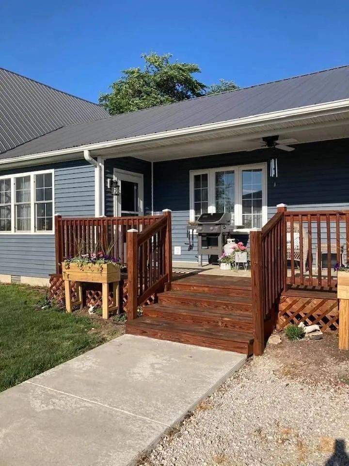 Front porch with wooden steps and railings on a gray house, beside a concrete walkway and gravel yard.