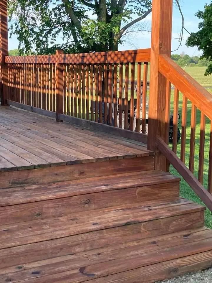 Wooden deck steps leading to a raised porch with red-brown railing and trees beyond