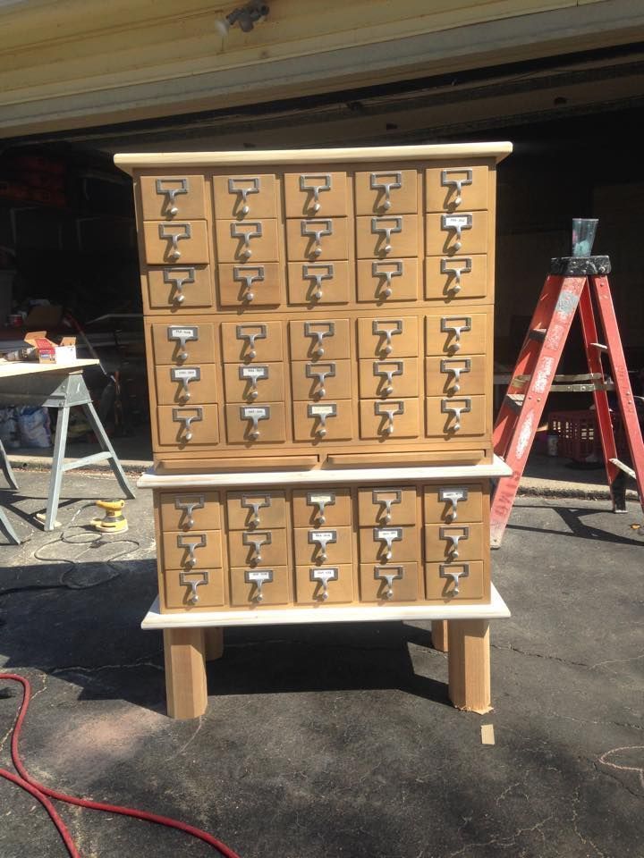 Wooden cabinet with many labeled drawers, standing on a driveway with a ladder and tools in the background.