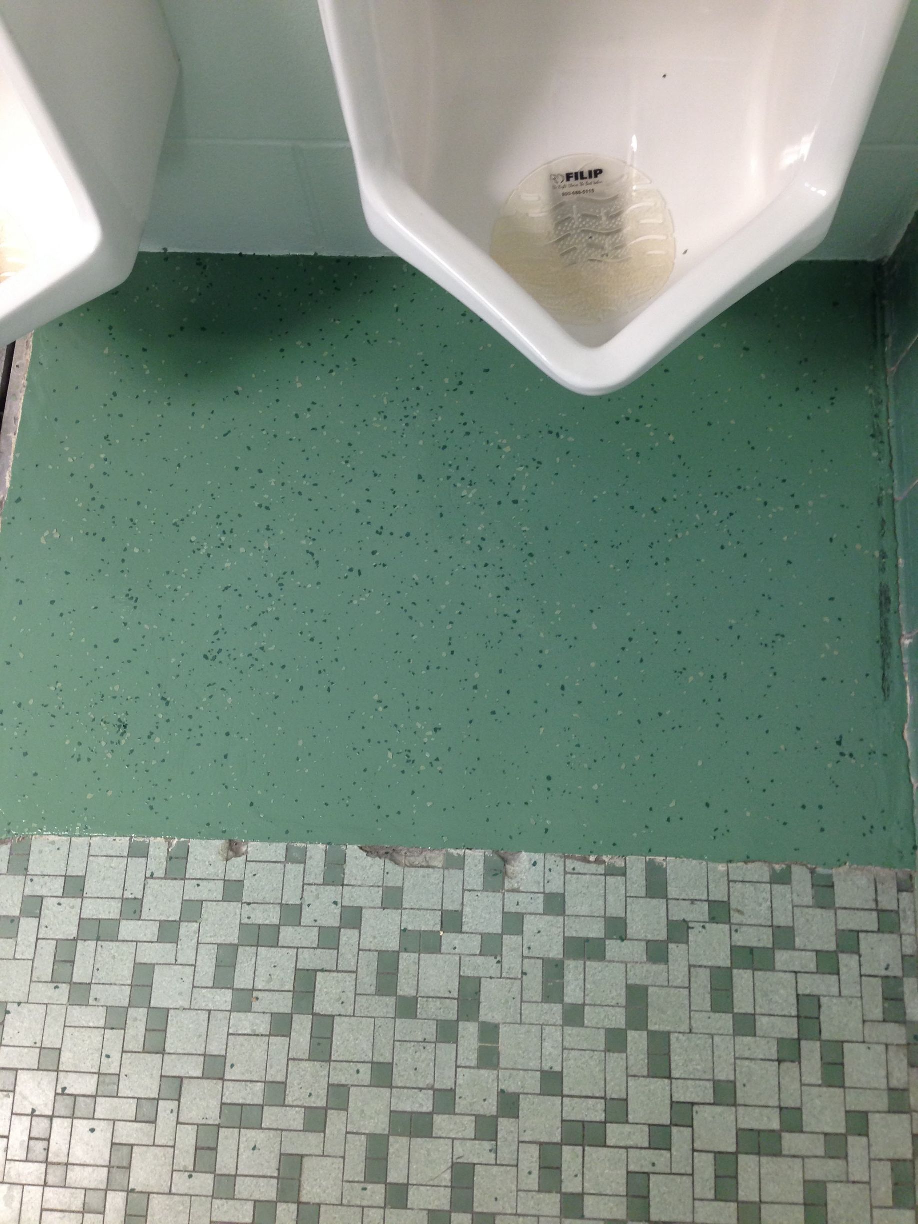 Public restroom with green speckled floor and white urinals against a tiled wall