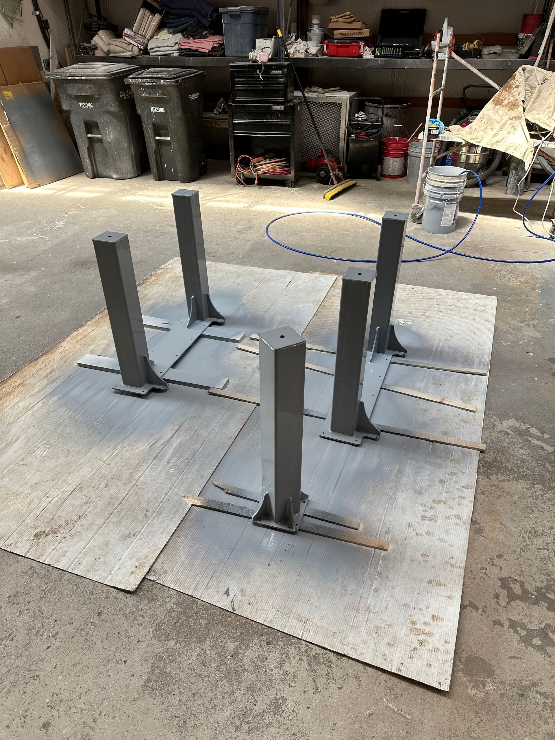 Four metal posts bolted to square plates on cardboard in a workshop floor area