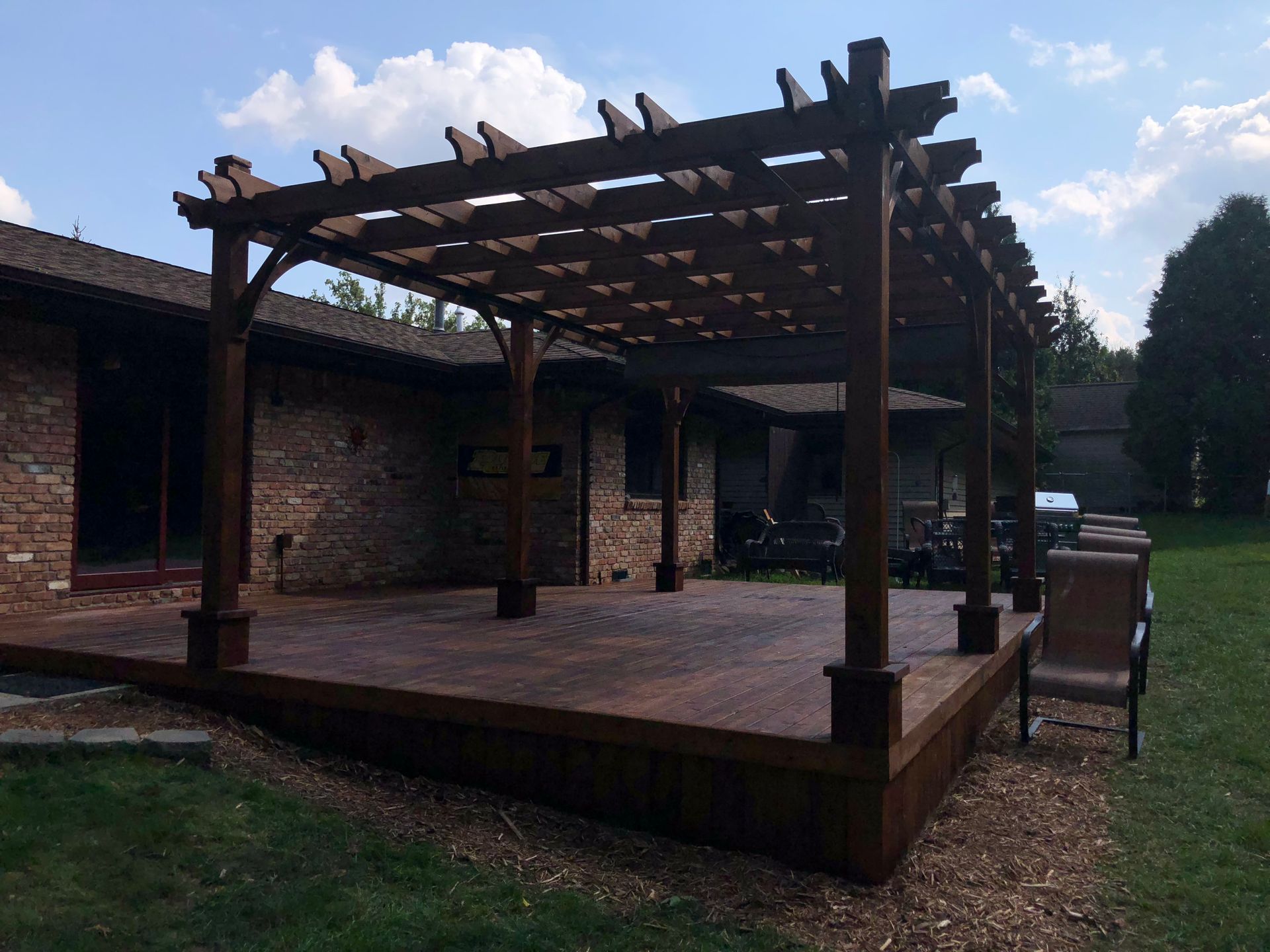 Wooden pergola-covered deck beside a brick building, with outdoor seating and a grassy yard.