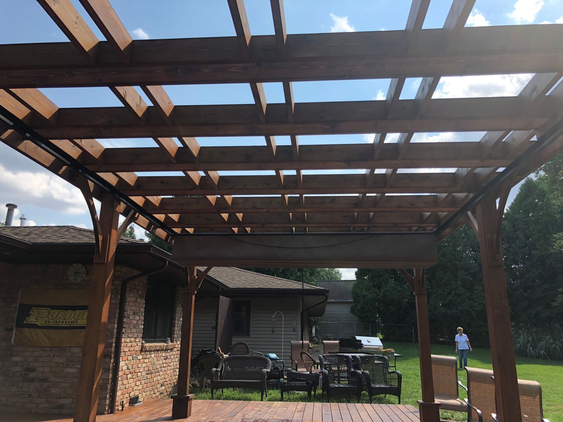 Wooden pergola over a patio beside a stone house, with outdoor furniture and a person in the background.