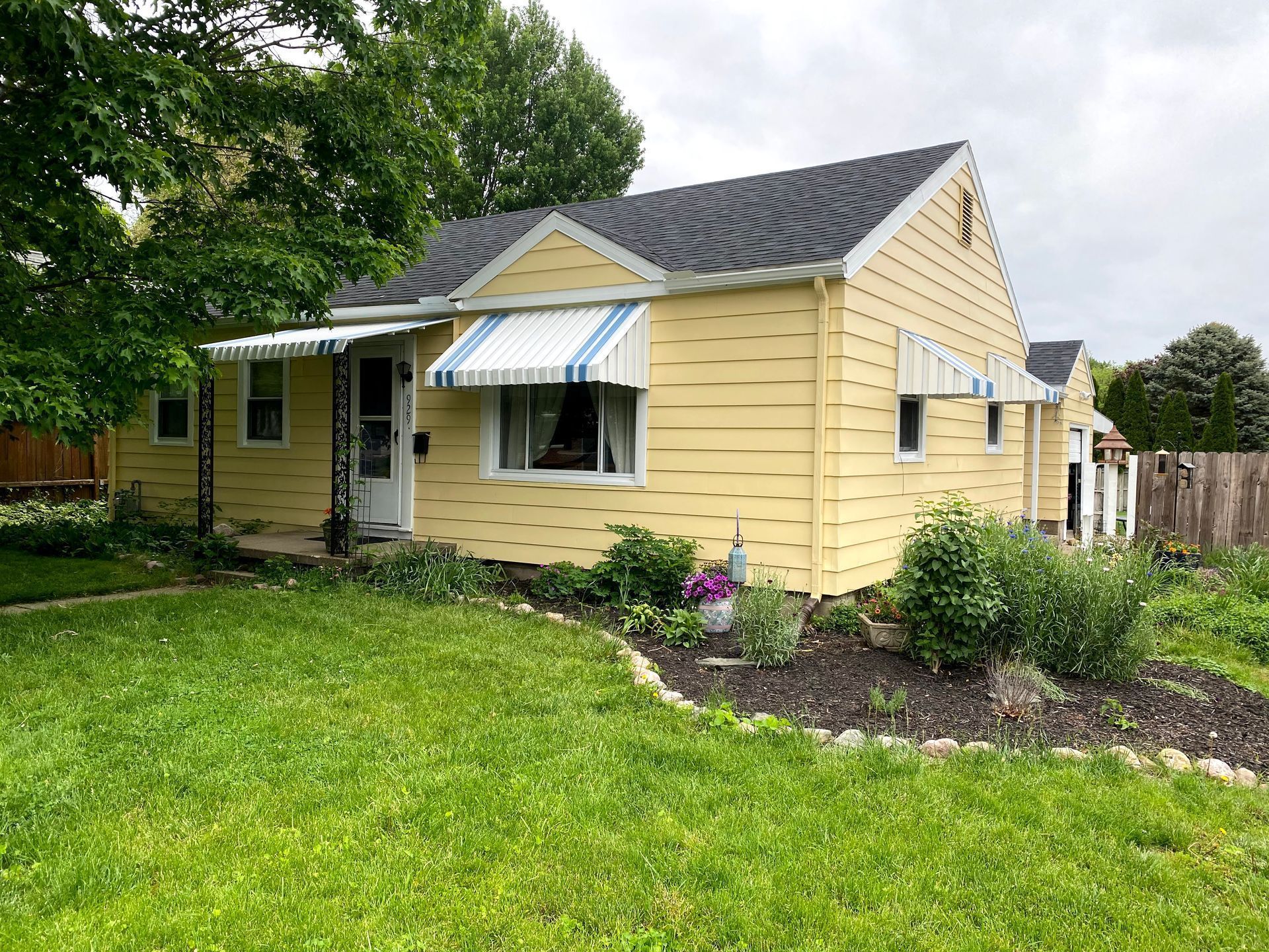 Small yellow house with striped awnings, a front porch, and a green yard with flower beds.