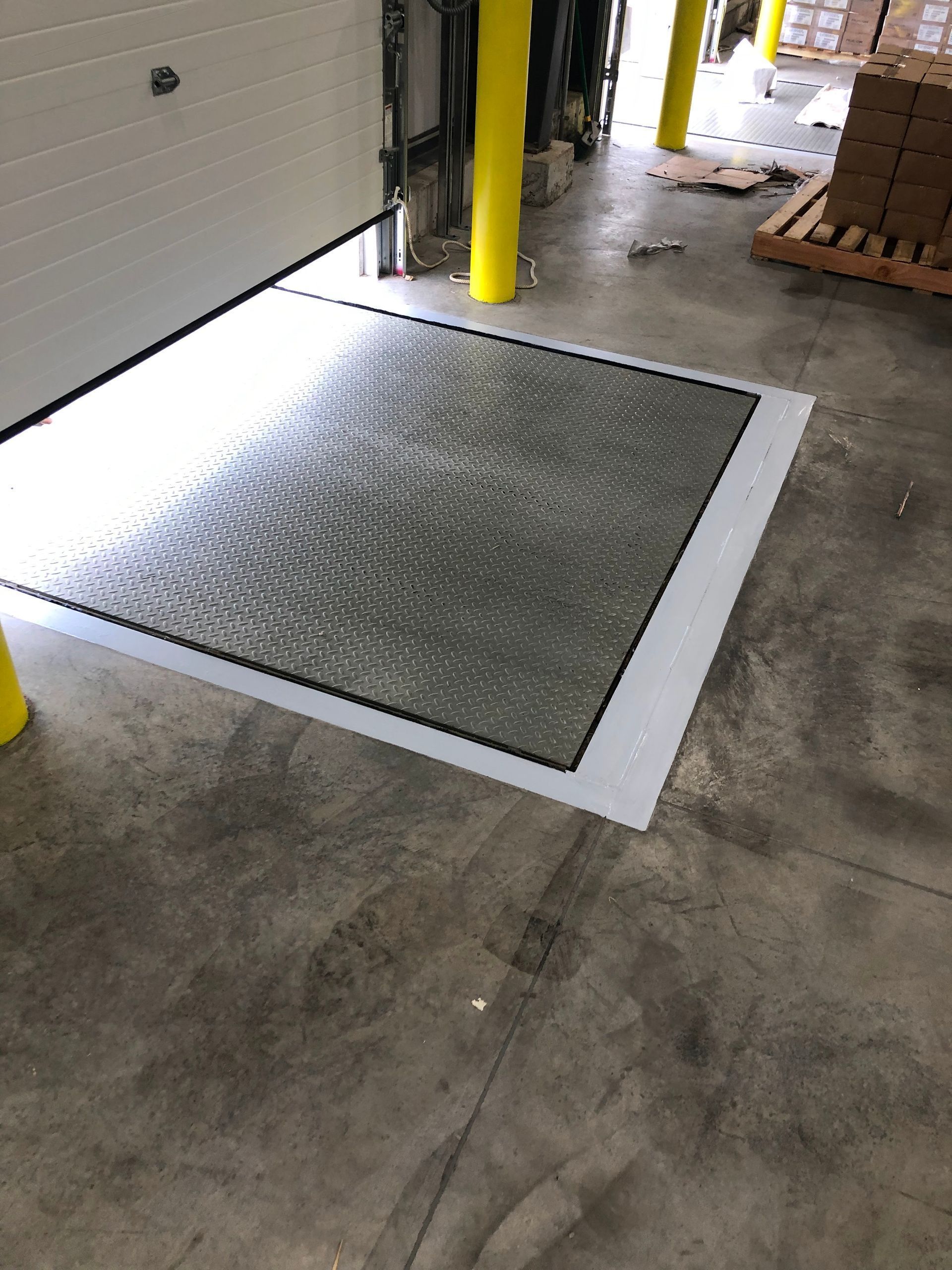 Metal floor mat beside a garage door, outlined in white on a concrete floor, with yellow safety posts nearby.