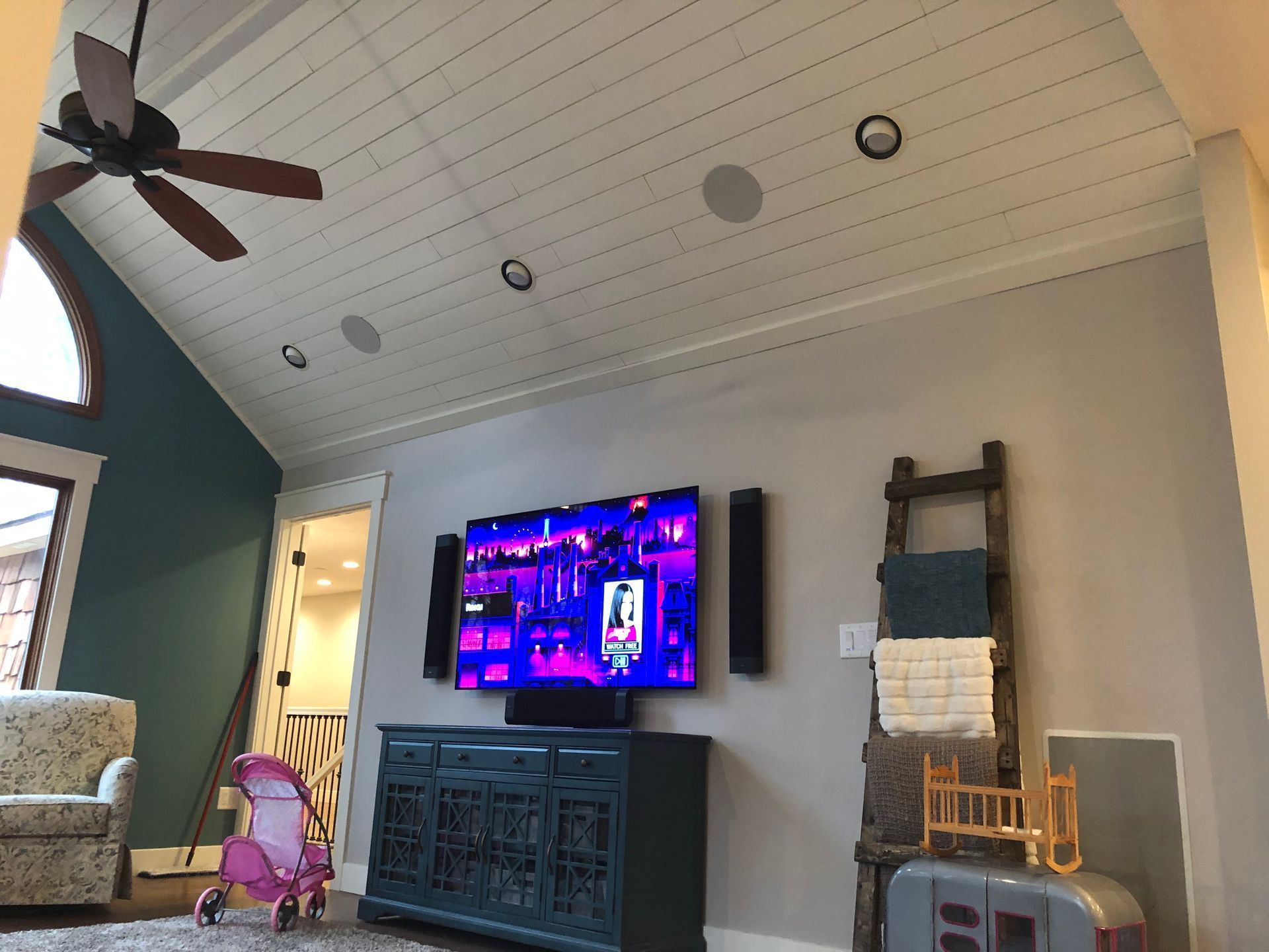 Living room with wall-mounted TV, ceiling fan, armchair, and a child’s toy stroller on the floor