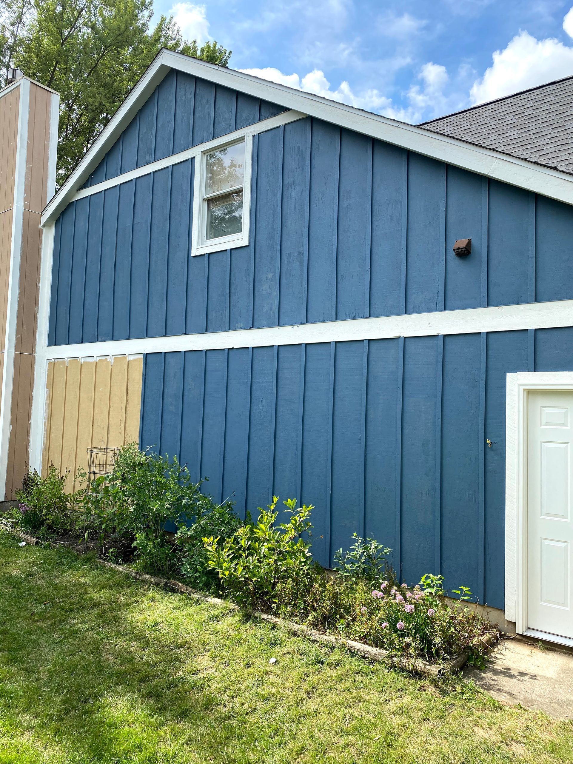 Blue house exterior with white trim, a small window, door, and patchy siding repair on the left.