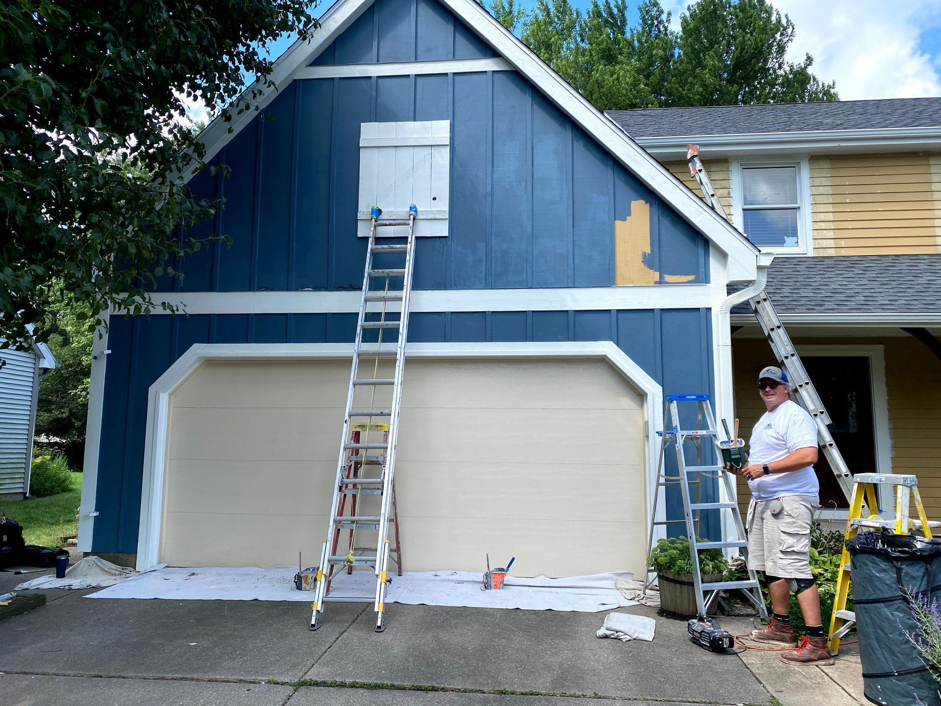 Worker on ladder painting blue house exterior above double garage.