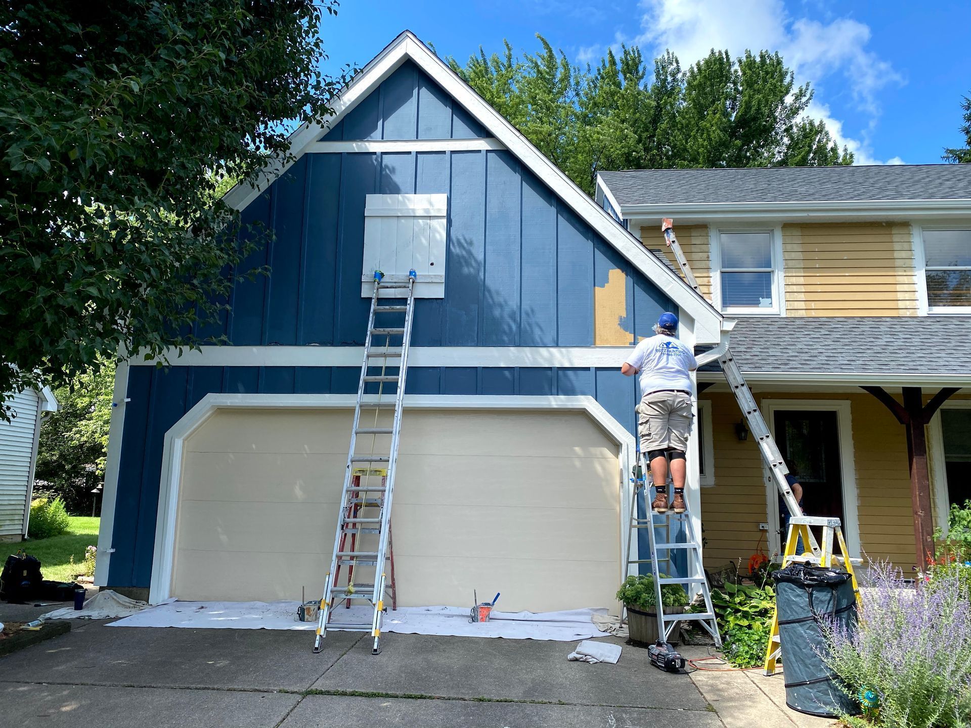 Workers painting a blue house exterior, with ladders set against the front and garage.