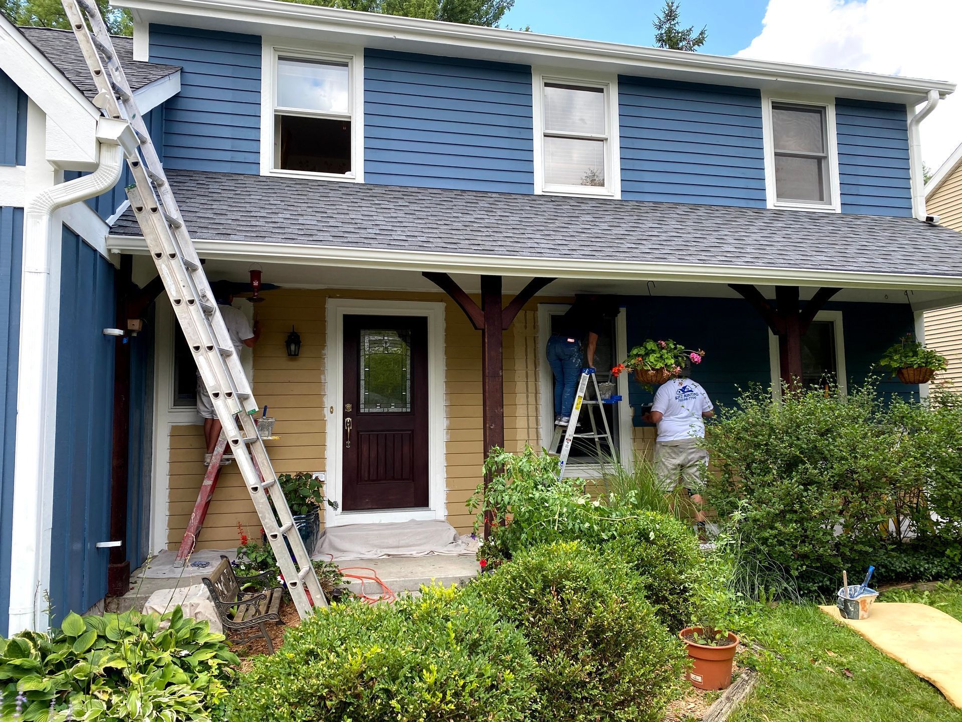 Blue two-story house with ladder leaning against it and people painting the porch.