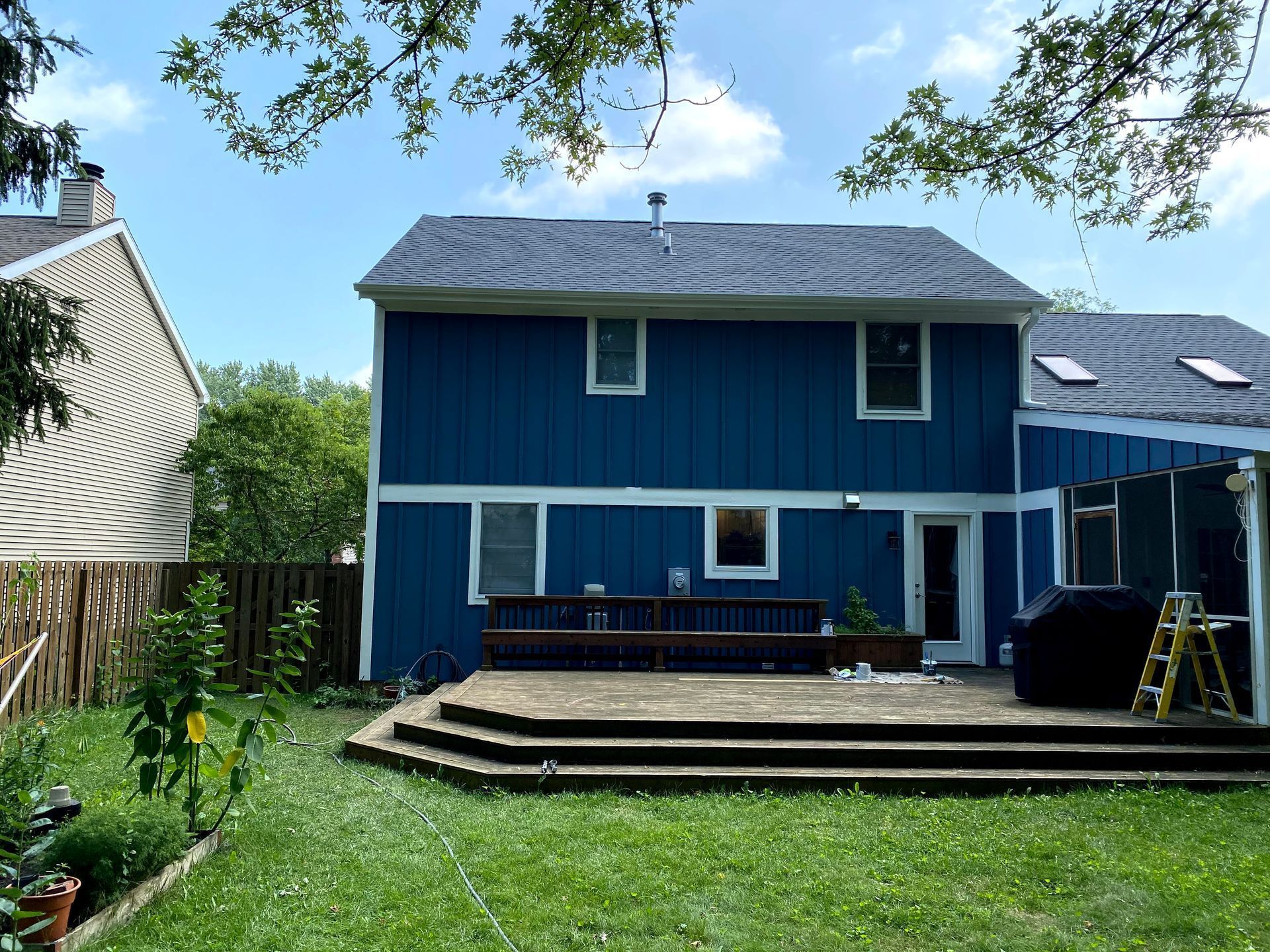 Blue two-story house with a wooden deck and green backyard under a partly cloudy sky