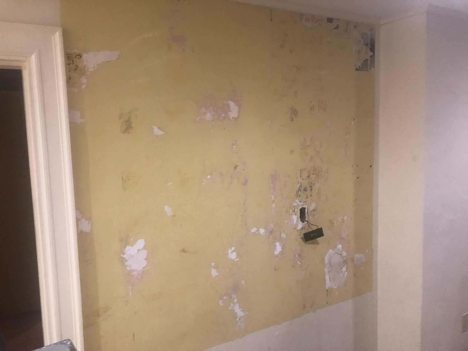Peeling yellow wall with patchy paint, exposed plaster, and an electrical outlet in a room corner