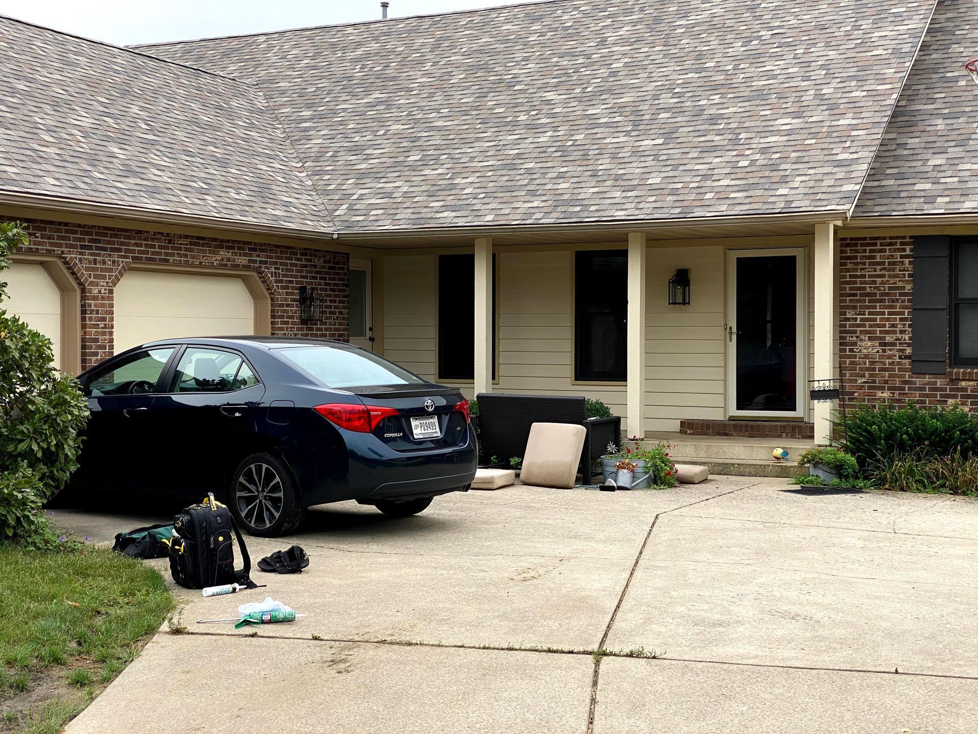 Black sedan parked in a driveway outside a brick suburban house, with bags on the ground.
