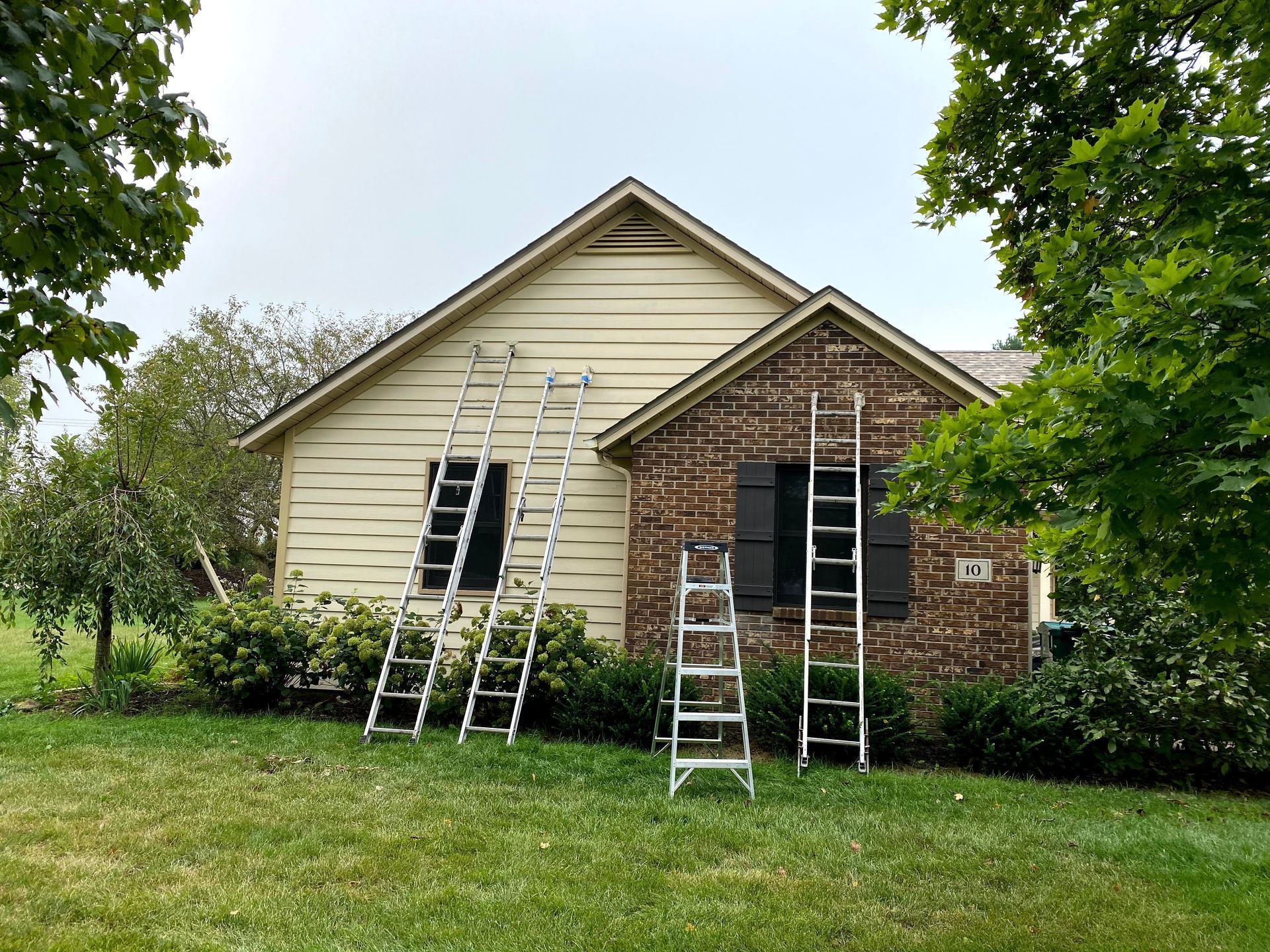 House exterior with ladders and scaffolding on the front, surrounded by green lawn and trees.