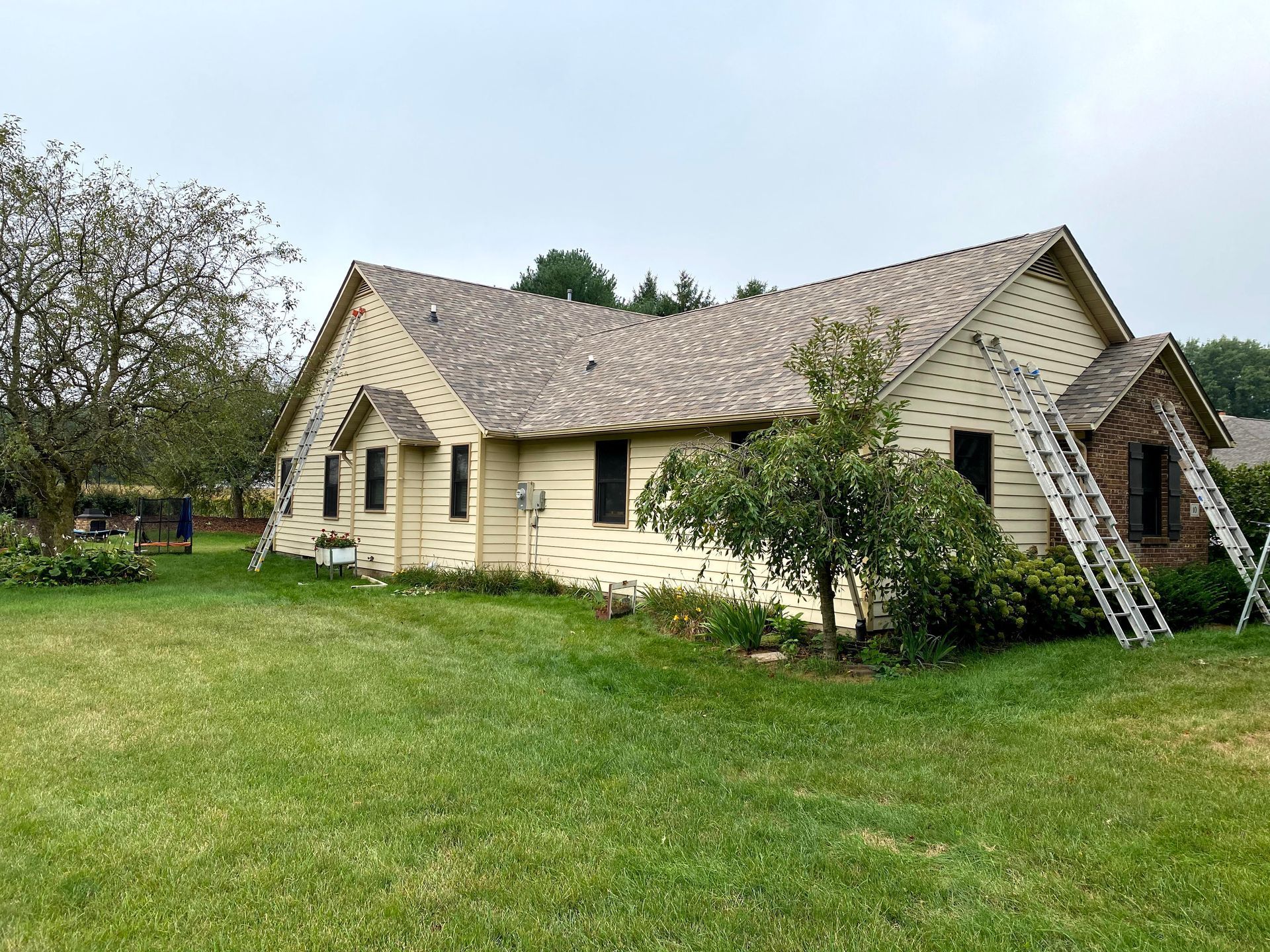 Single-story beige house with a steep roof and ladder leaning against it, on a grassy yard.