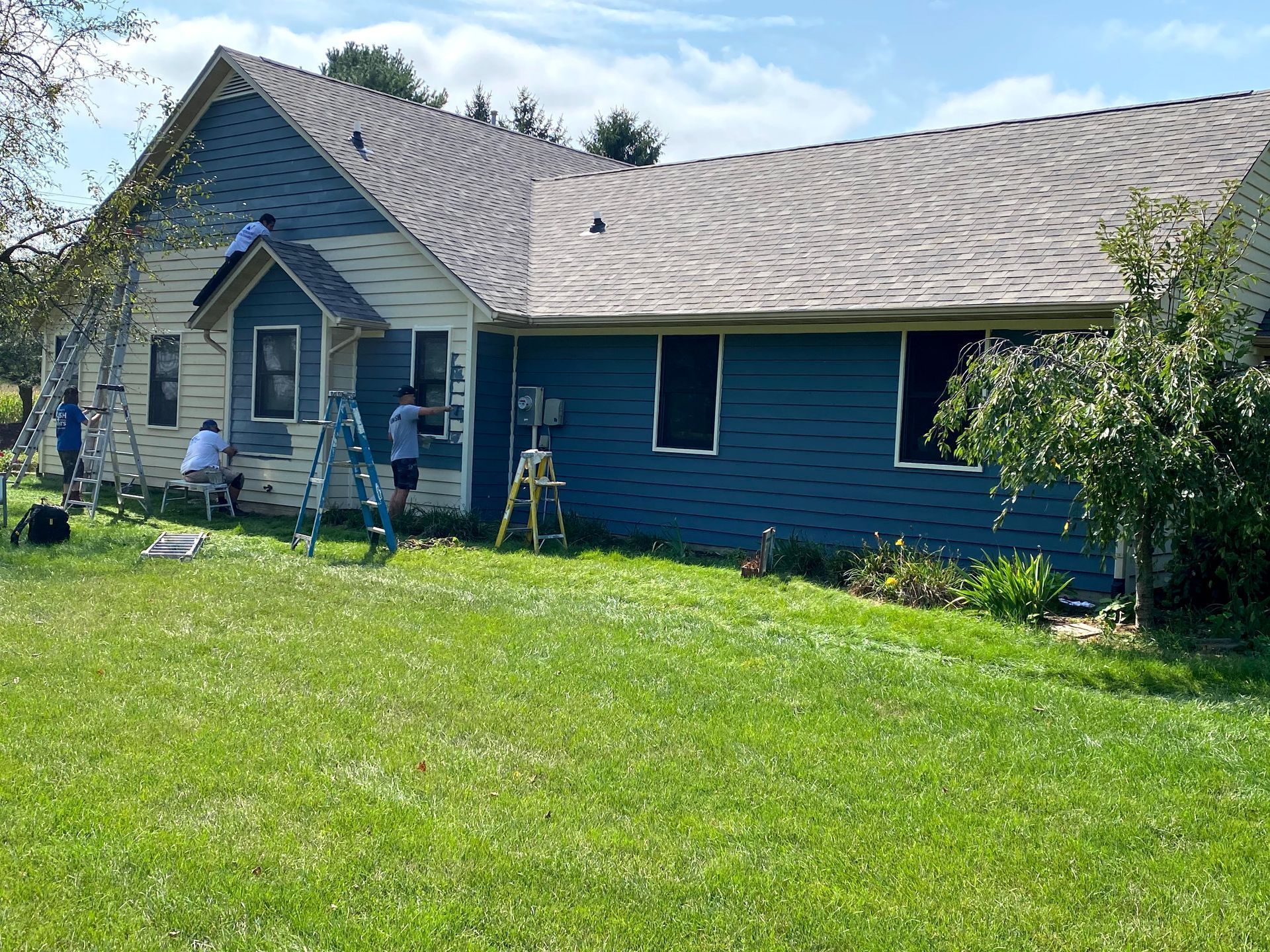 Blue house with white trim and a front porch, with people working in the yard on a sunny day