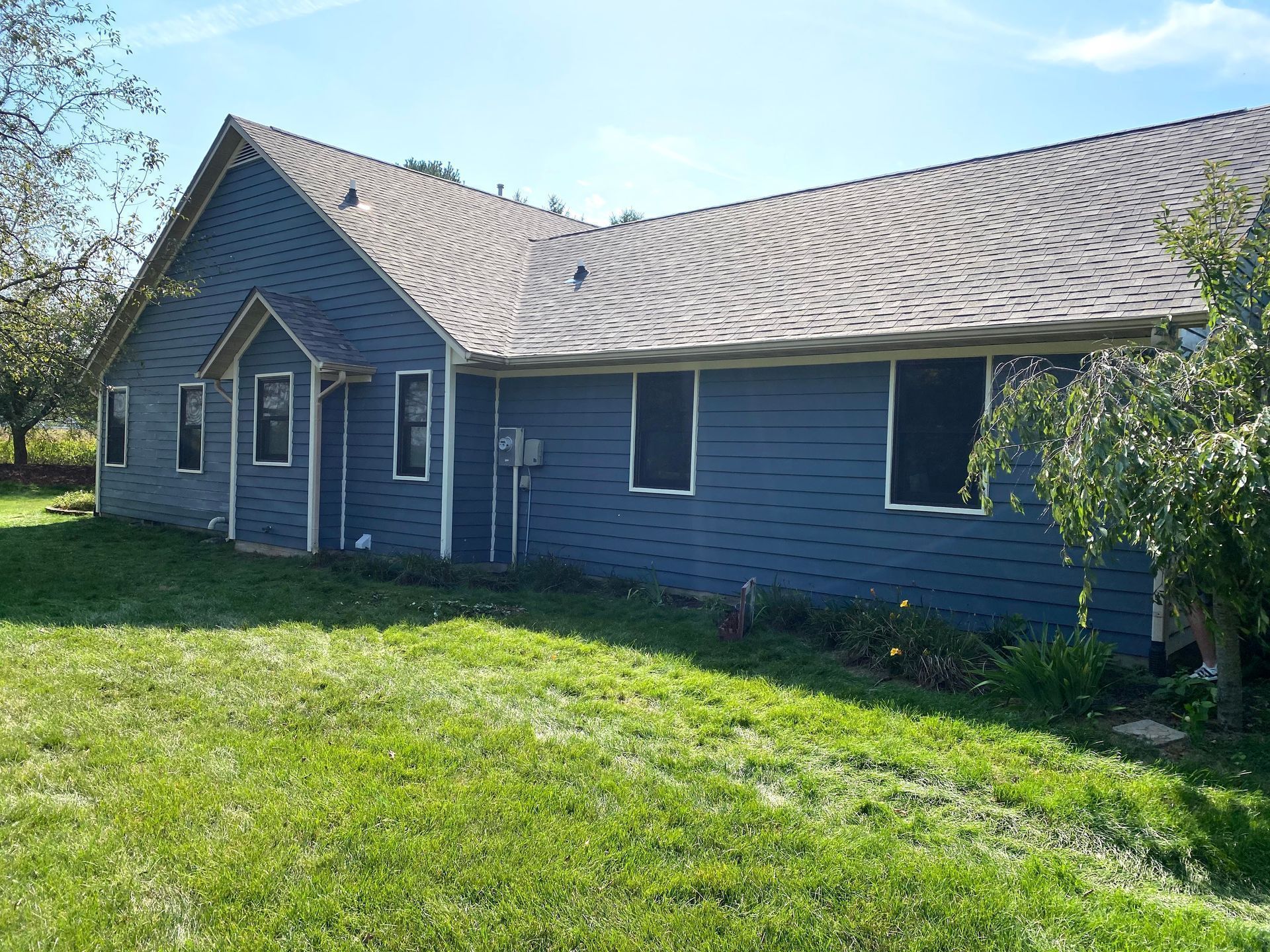 Blue house with a gray roof and white-trimmed windows, standing beside a grassy yard on a sunny day