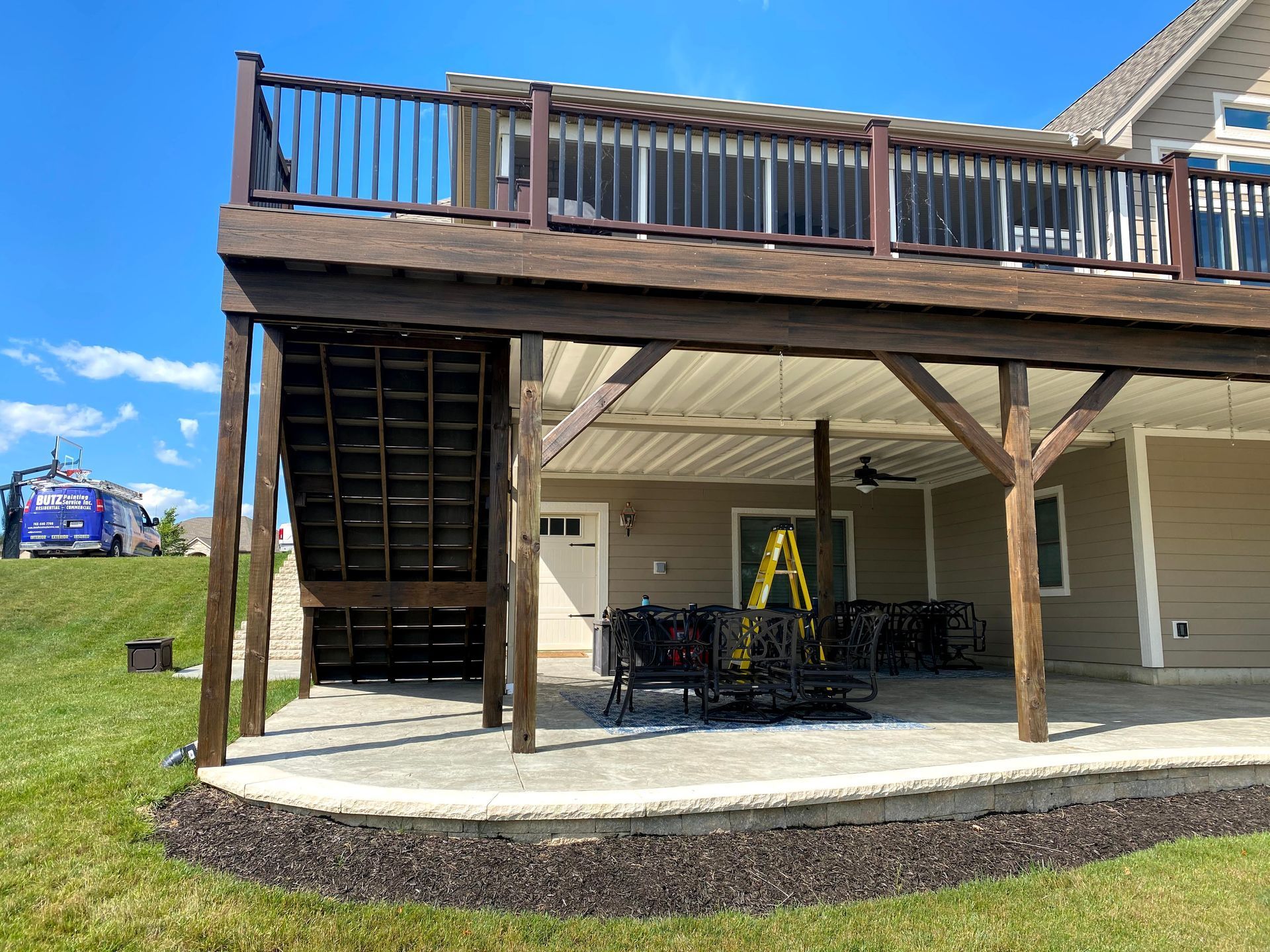 Covered patio under a wooden deck with stairs, railing, and an A-frame sign outside a beige building.
