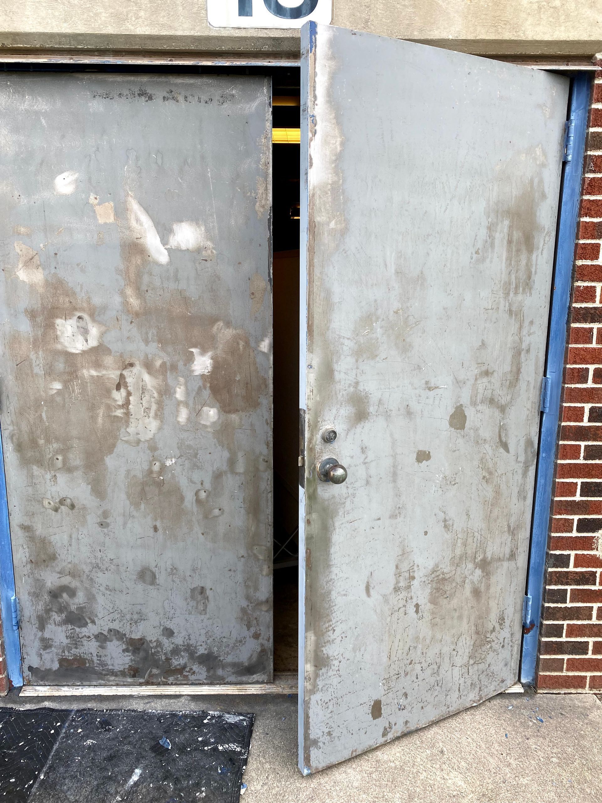 Open metal door on a worn gray industrial building, with blue trim and brick wall beside it.