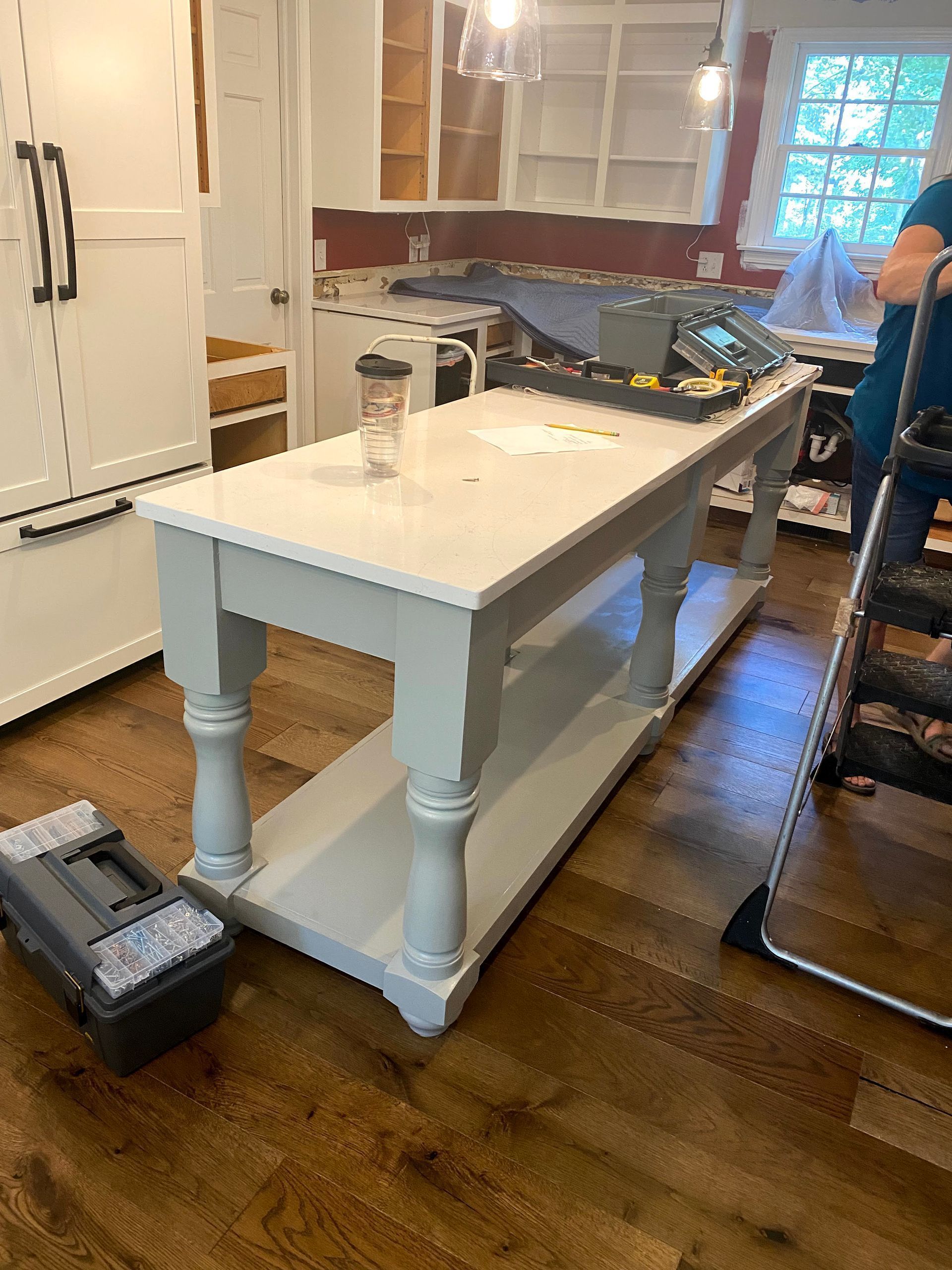 White kitchen island on hardwood floor during renovation, with tools and a worker partially visible on the right.