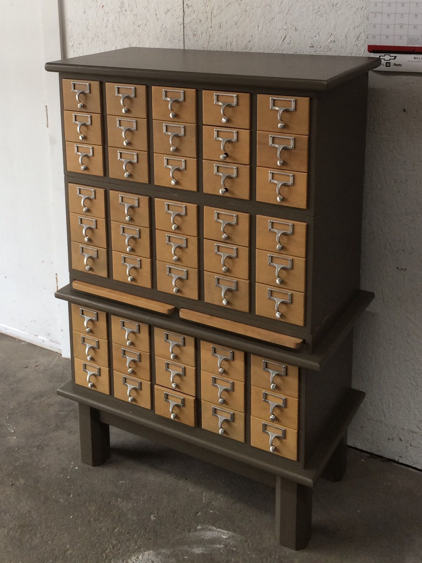 Antique wooden card catalog cabinet with many small drawers, standing against a speckled wall.