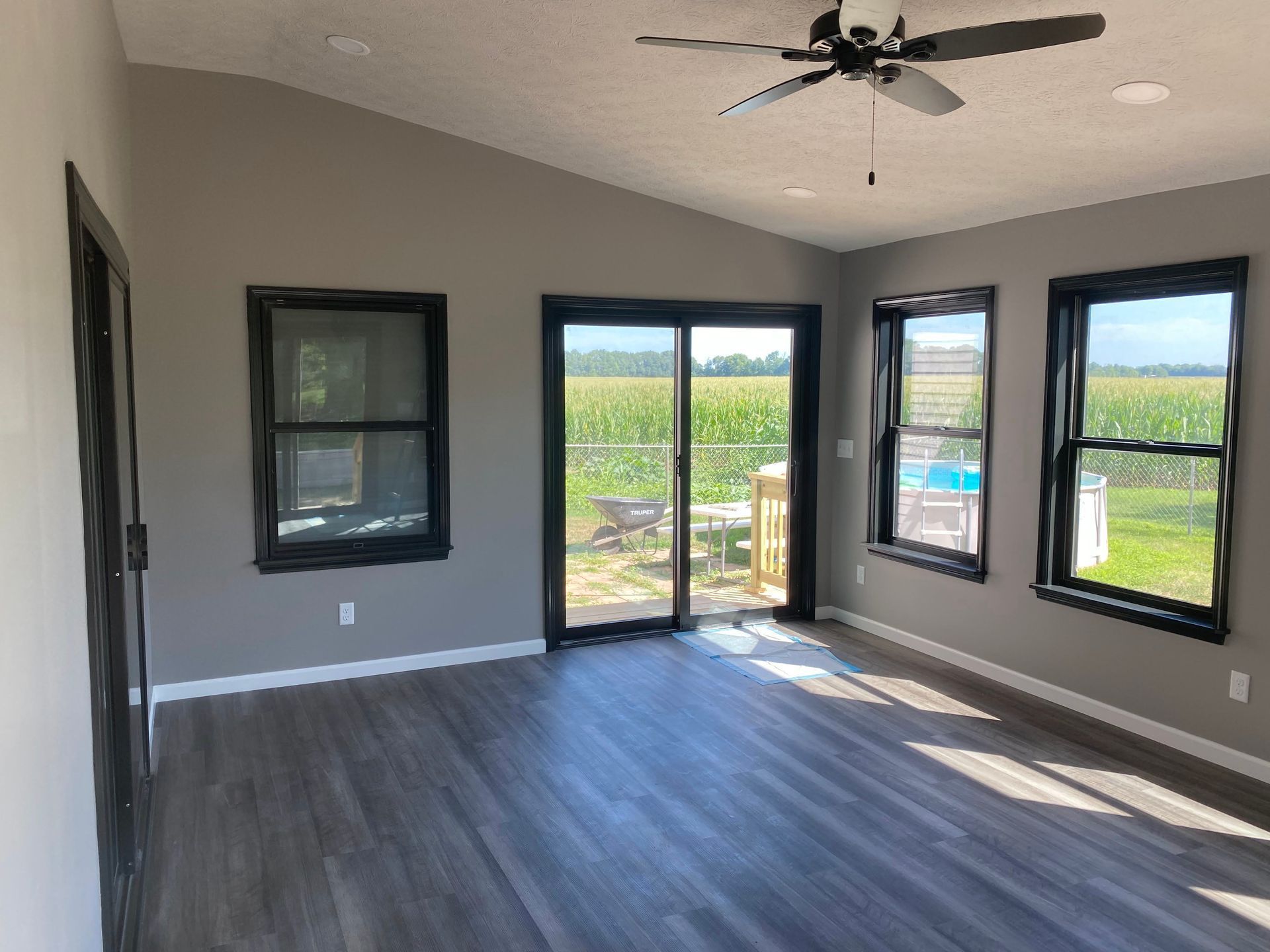 Empty modern room with dark wood floor, ceiling fan, and large windows overlooking a green field