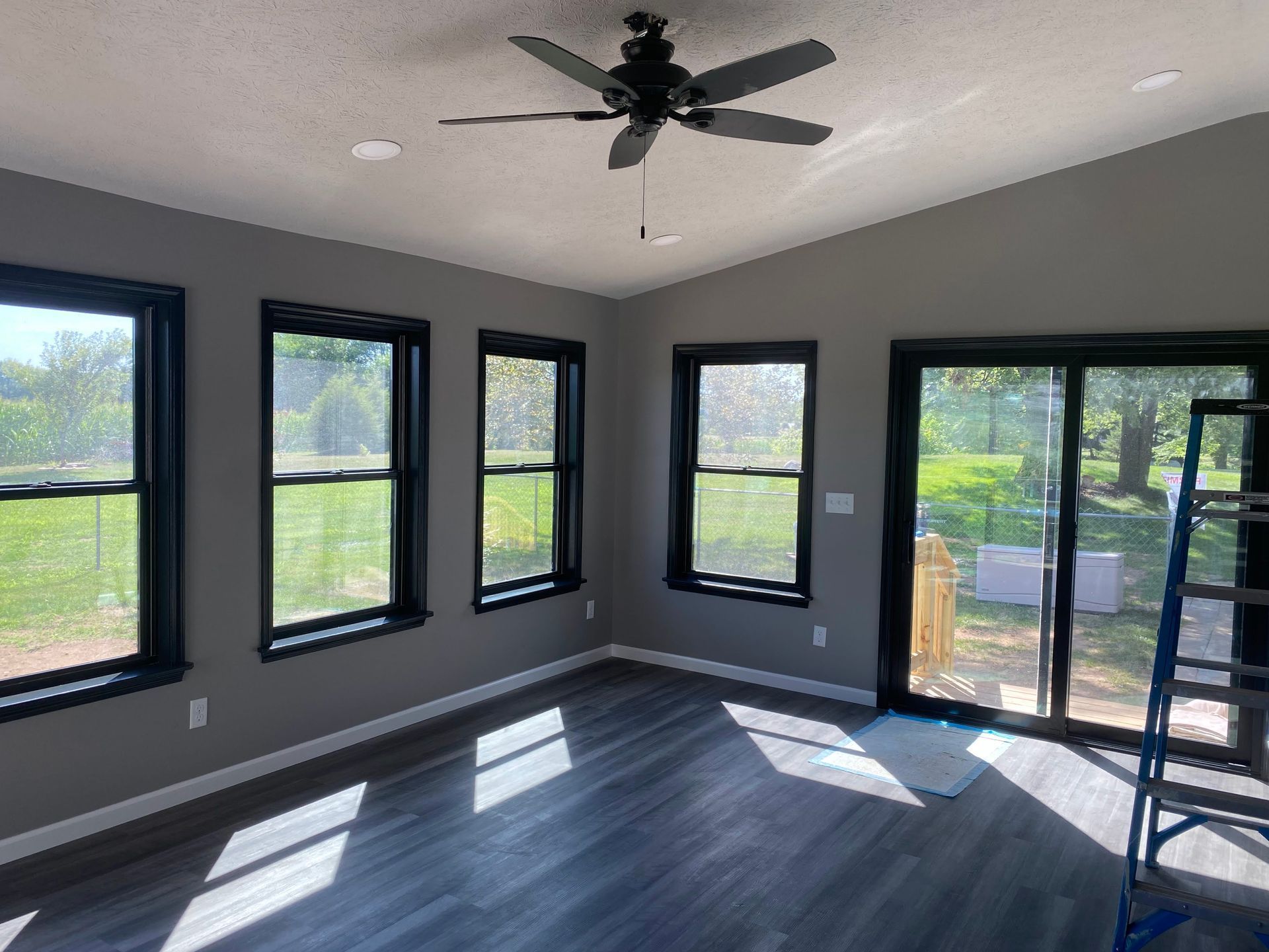 Empty gray room with black-trim windows, ceiling fan, sliding glass door, and ladder by the wall