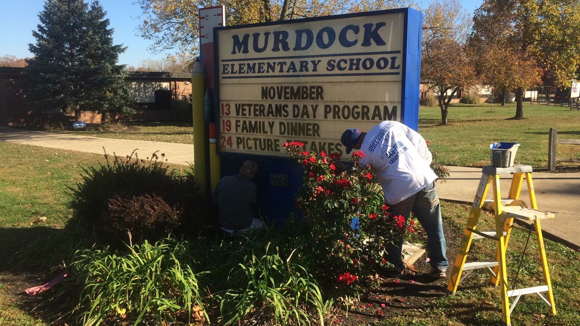 Person trims flowers beside the Murdock Elementary School sign on a sunny day.