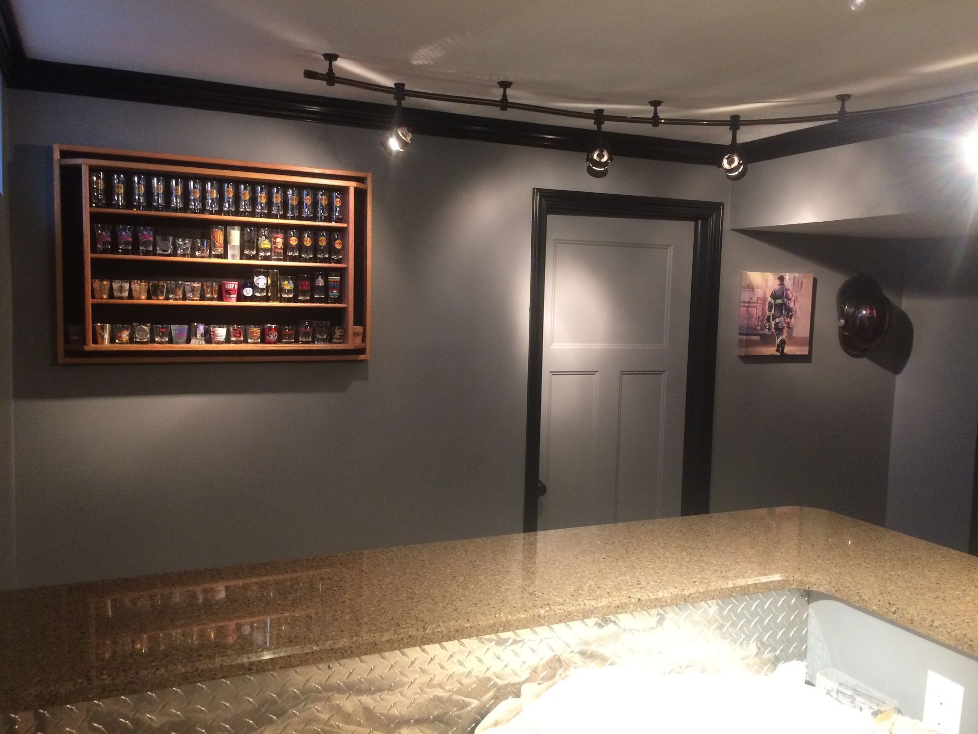 Dim home bar with a granite counter, wall-mounted liquor shelf, closed door, and track lighting.