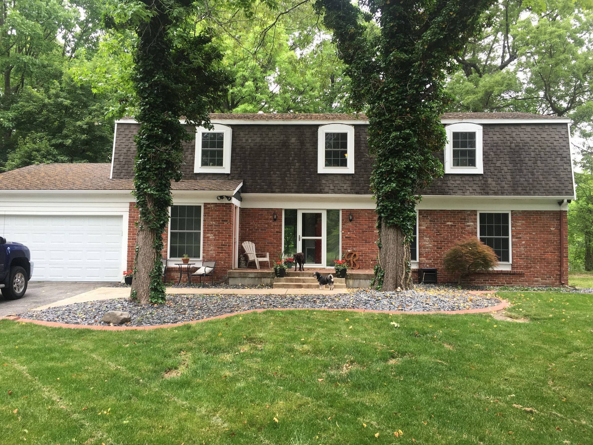 Suburban brick house with white garage, dormer windows, and a front lawn framed by tall trees