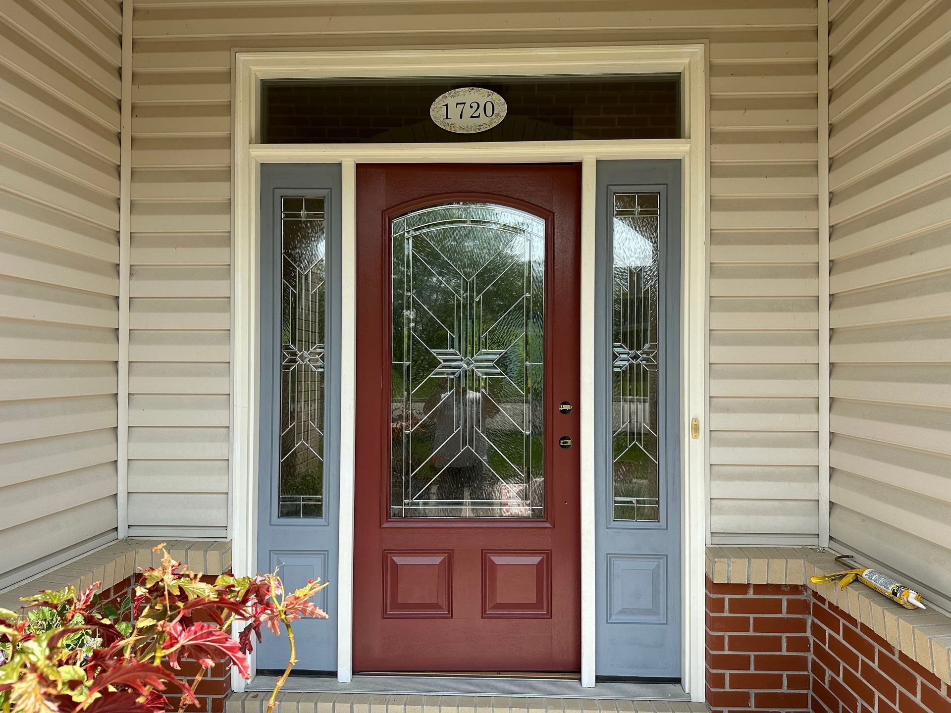 Front porch with a red front door, sidelights, and brick steps, framed by cream siding and potted plants