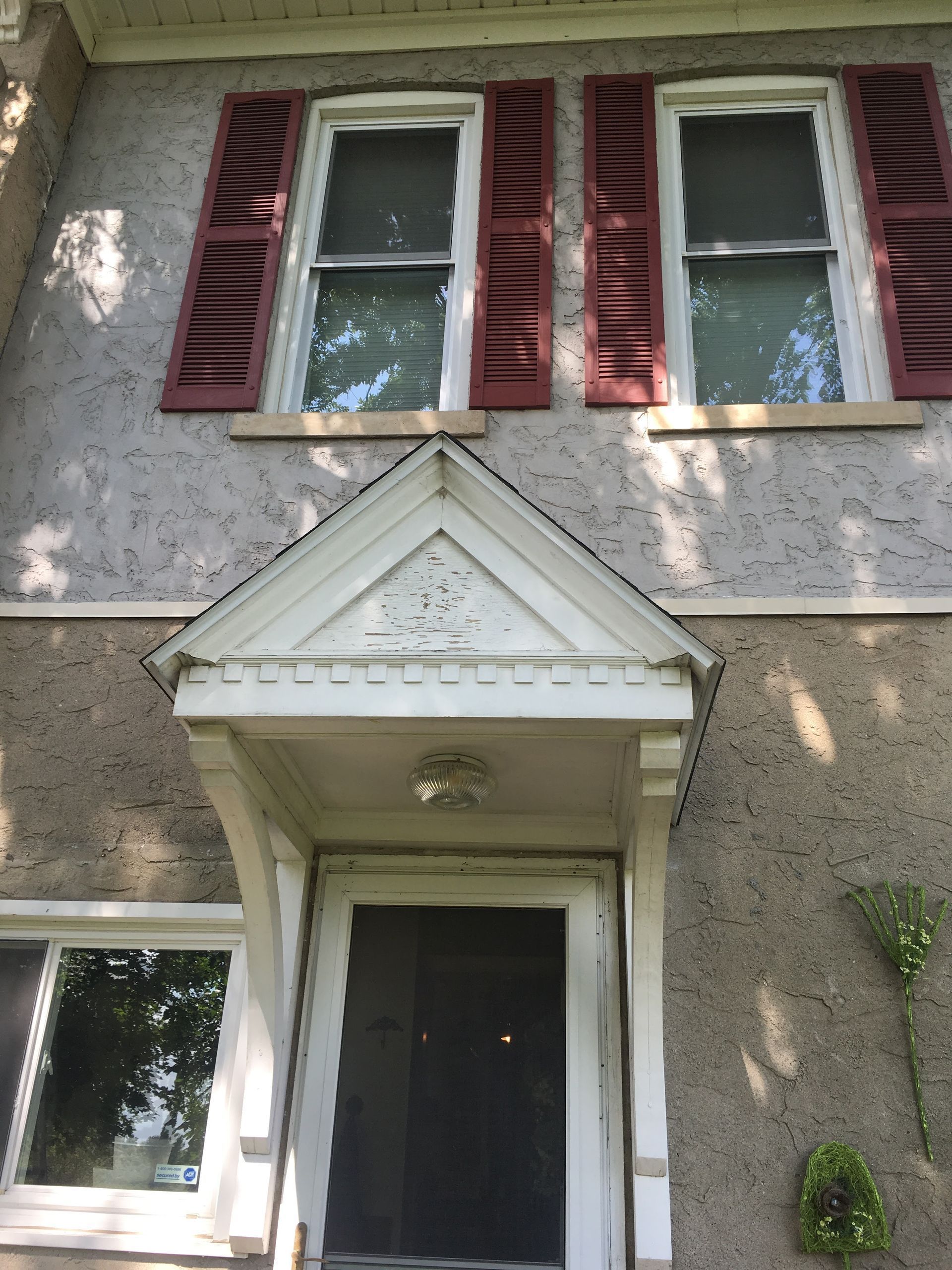 Front entrance of a stucco house with red shutters, white trim, and a small peaked porch roof.