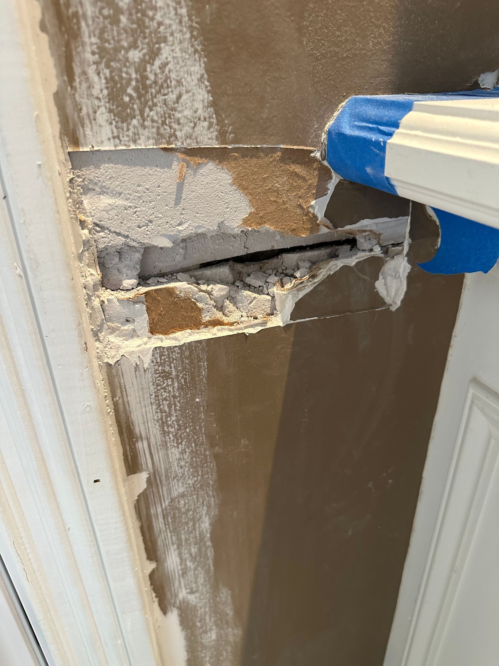 Damaged wall corner with a large hole and peeling paint near blue painter’s tape