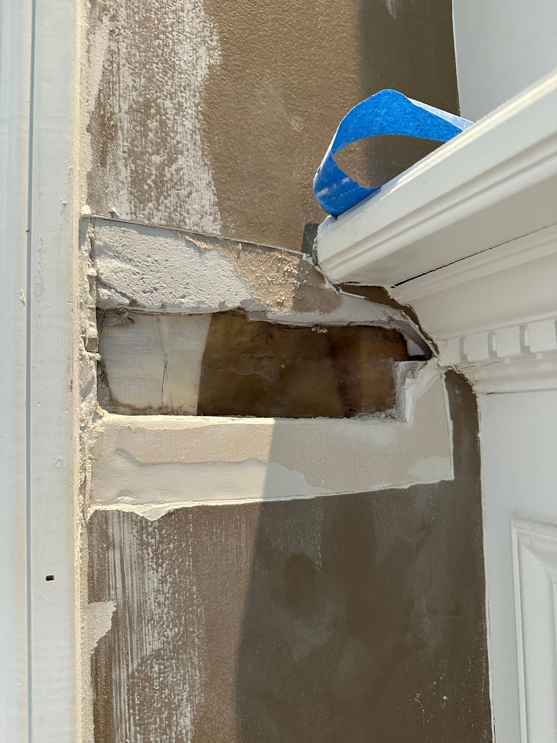 Damaged wall opening beside white trim, with exposed drywall and blue tape on top
