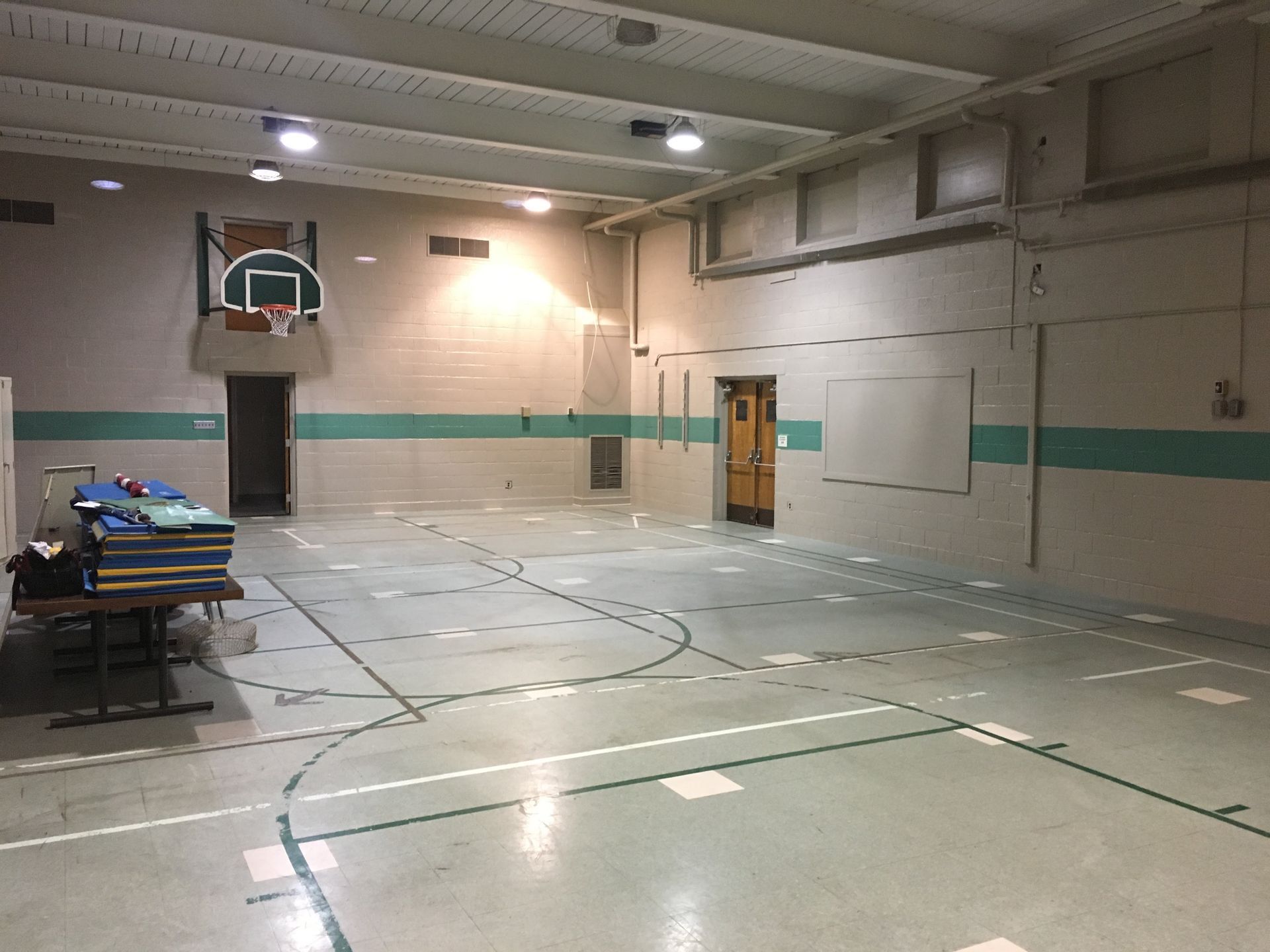 Empty indoor gymnasium with a basketball hoop, folding table, and taped court lines