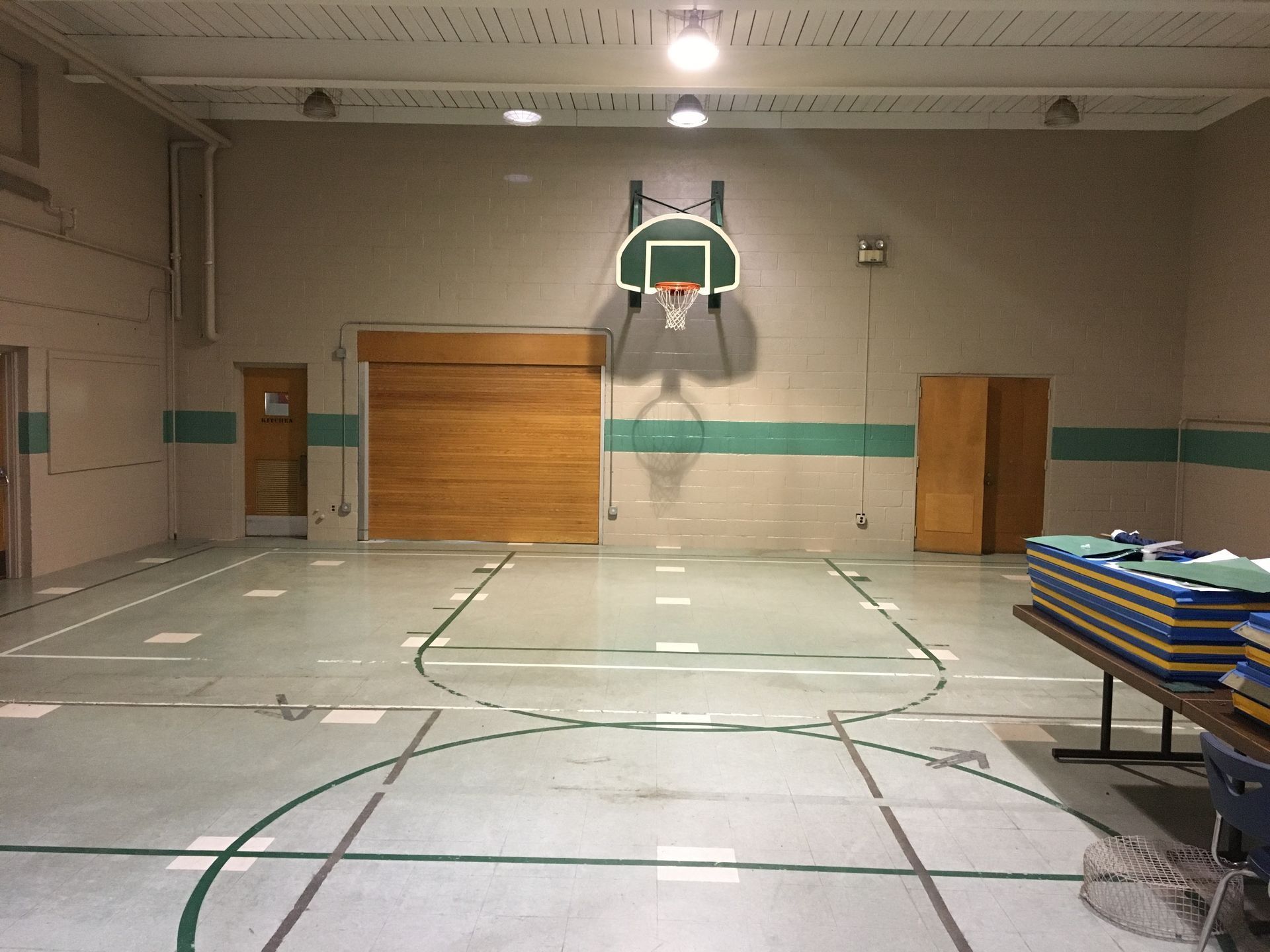 Empty school gym with a basketball hoop and court lines, wooden wall panels, and stacked mats on the right