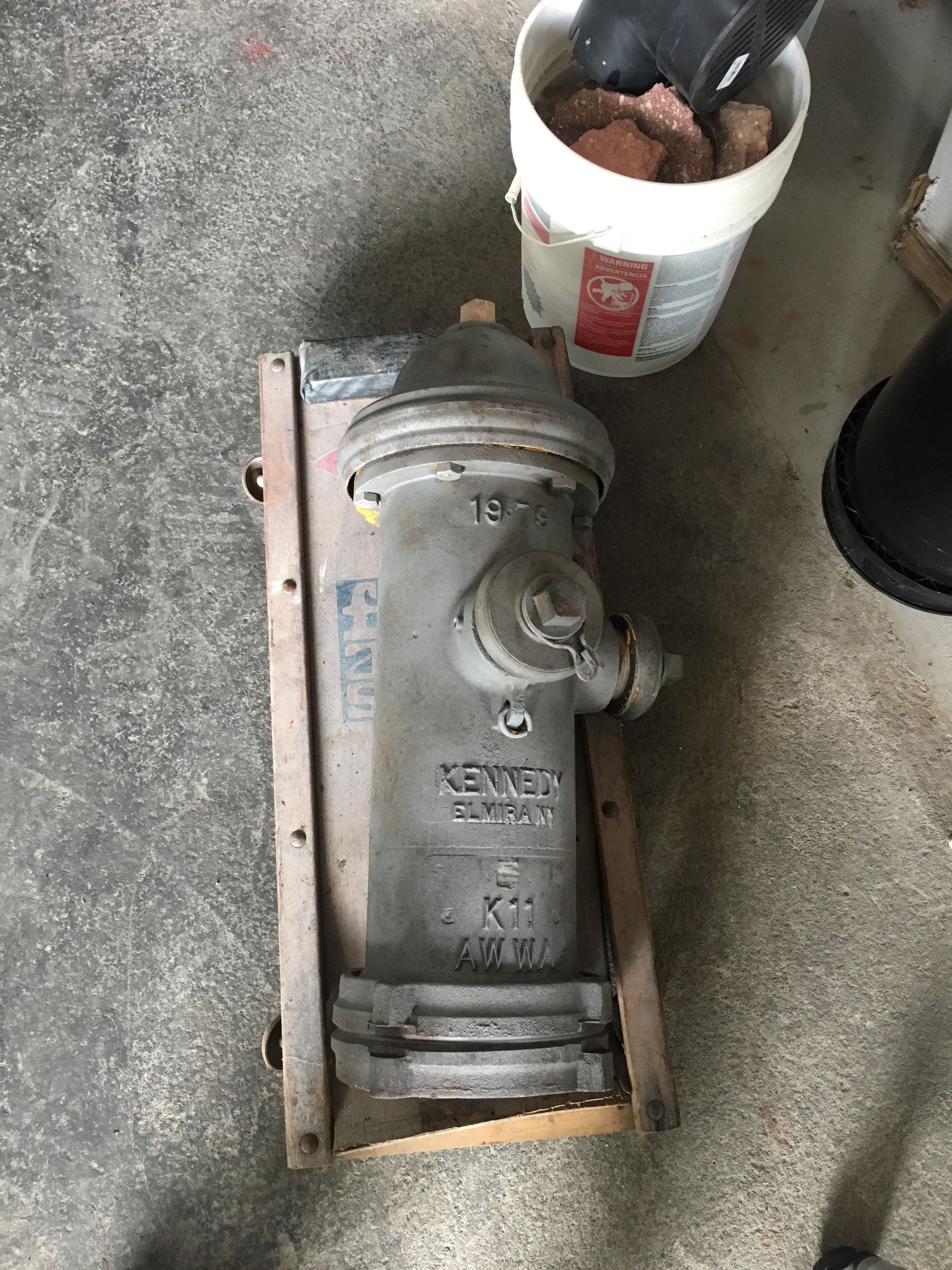 Gray metal fire hydrant or valve on a wooden pallet beside a bucket of tools on a concrete floor