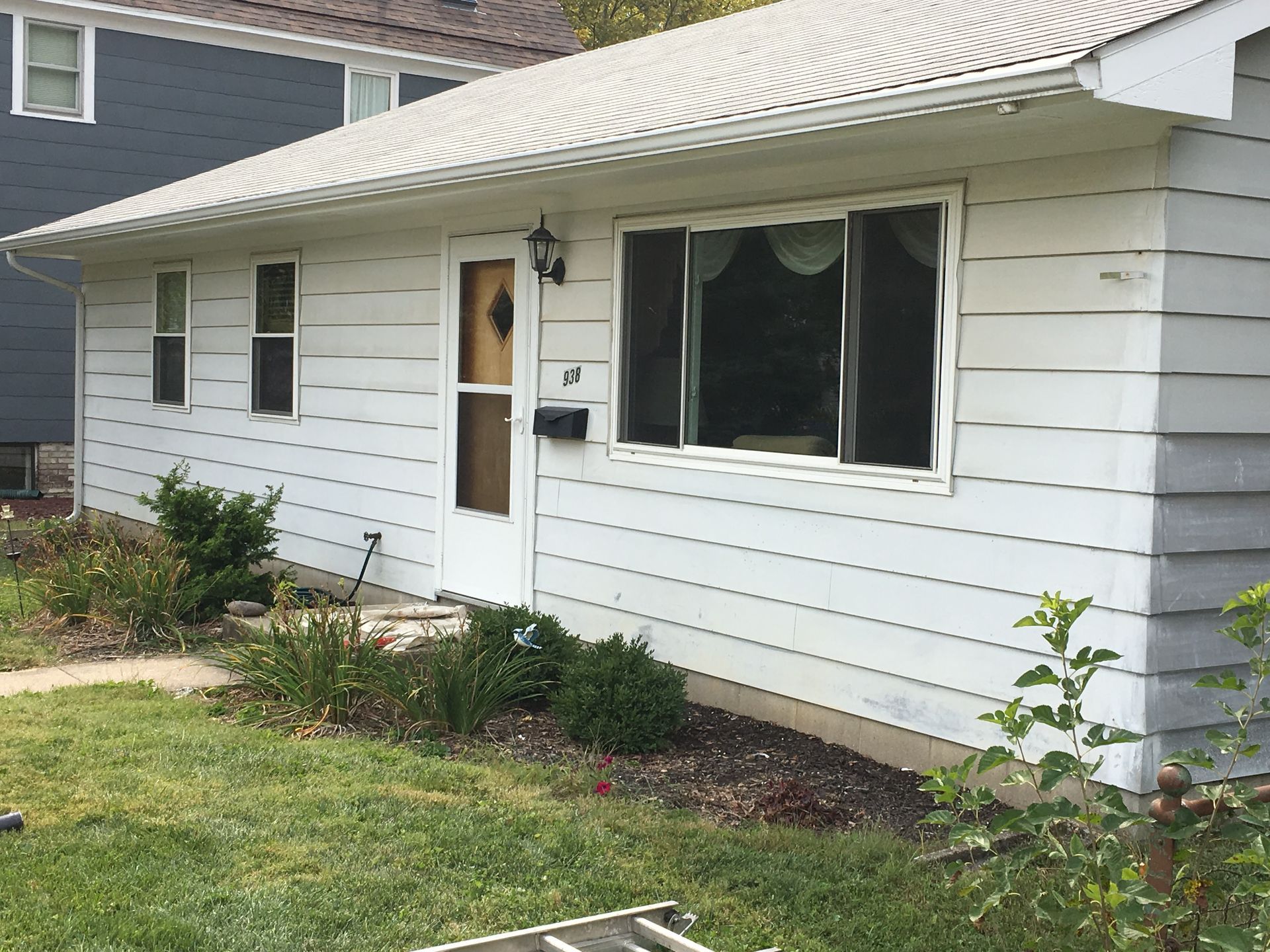 White single-story house exterior with siding, front door, windows, and small lawn and shrubs