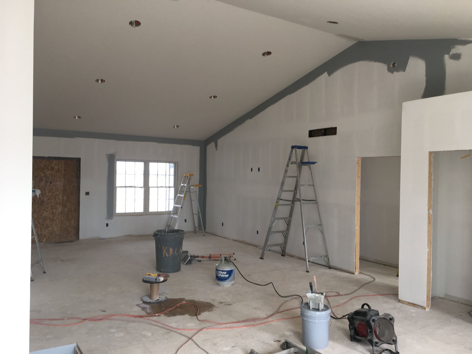 Unfinished room under renovation with ladders, tools, paint buckets, and exposed drywall.
