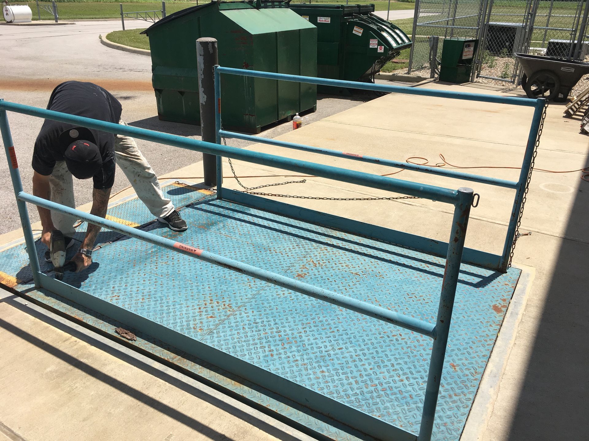 Worker sanding a blue metal platform beside green dumpsters outdoors