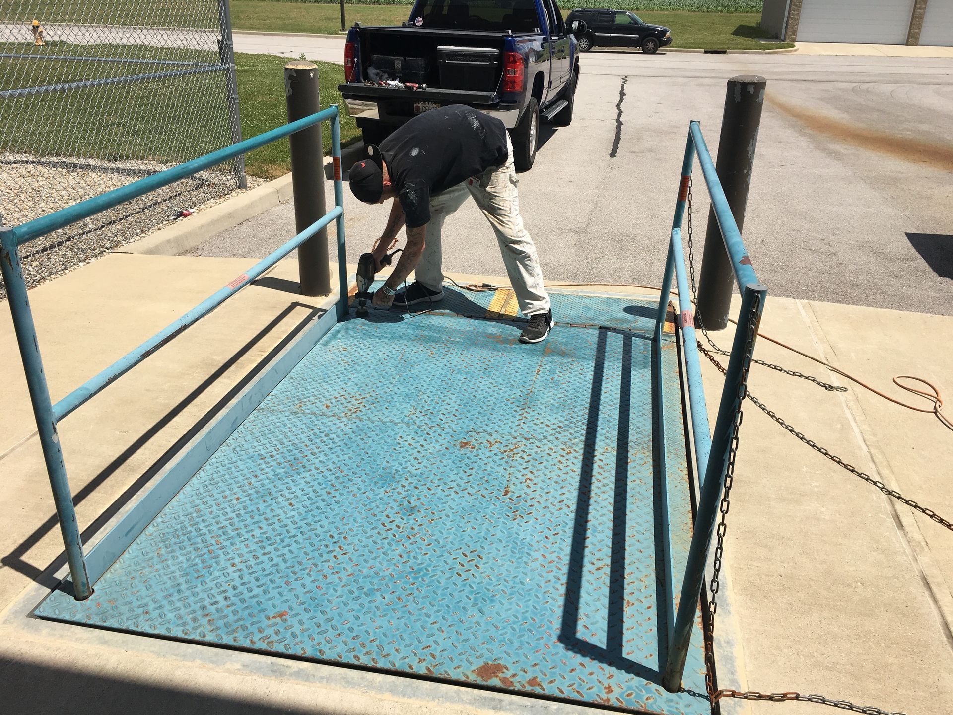 Person welding a blue metal ramp beside a black pickup truck on a sunny driveway.