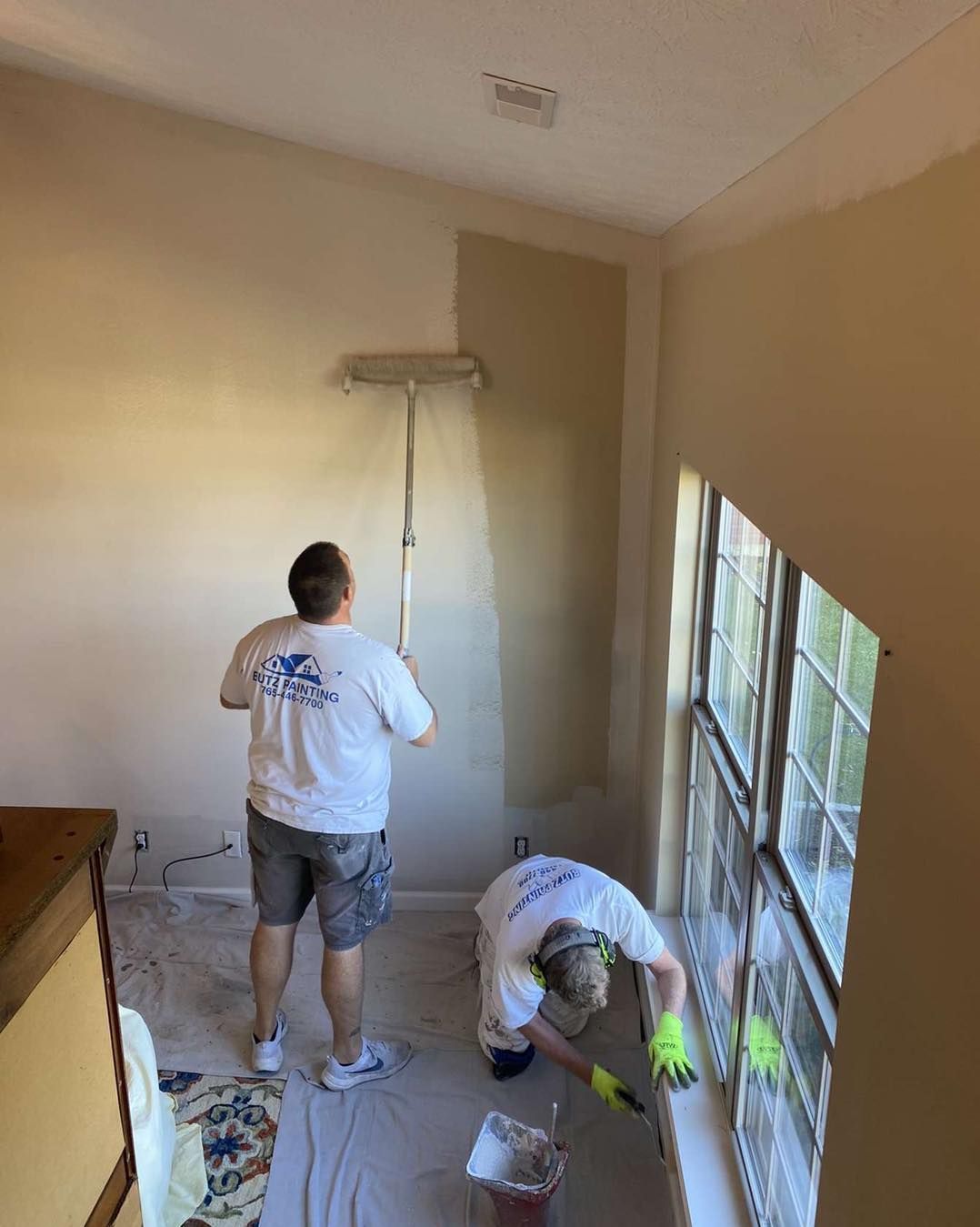 Two people painting a beige room, one on a ladder near a window and one crouching by supplies.
