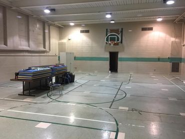 Empty indoor gymnasium with a basketball hoop, folded bleachers, and court lines on the floor