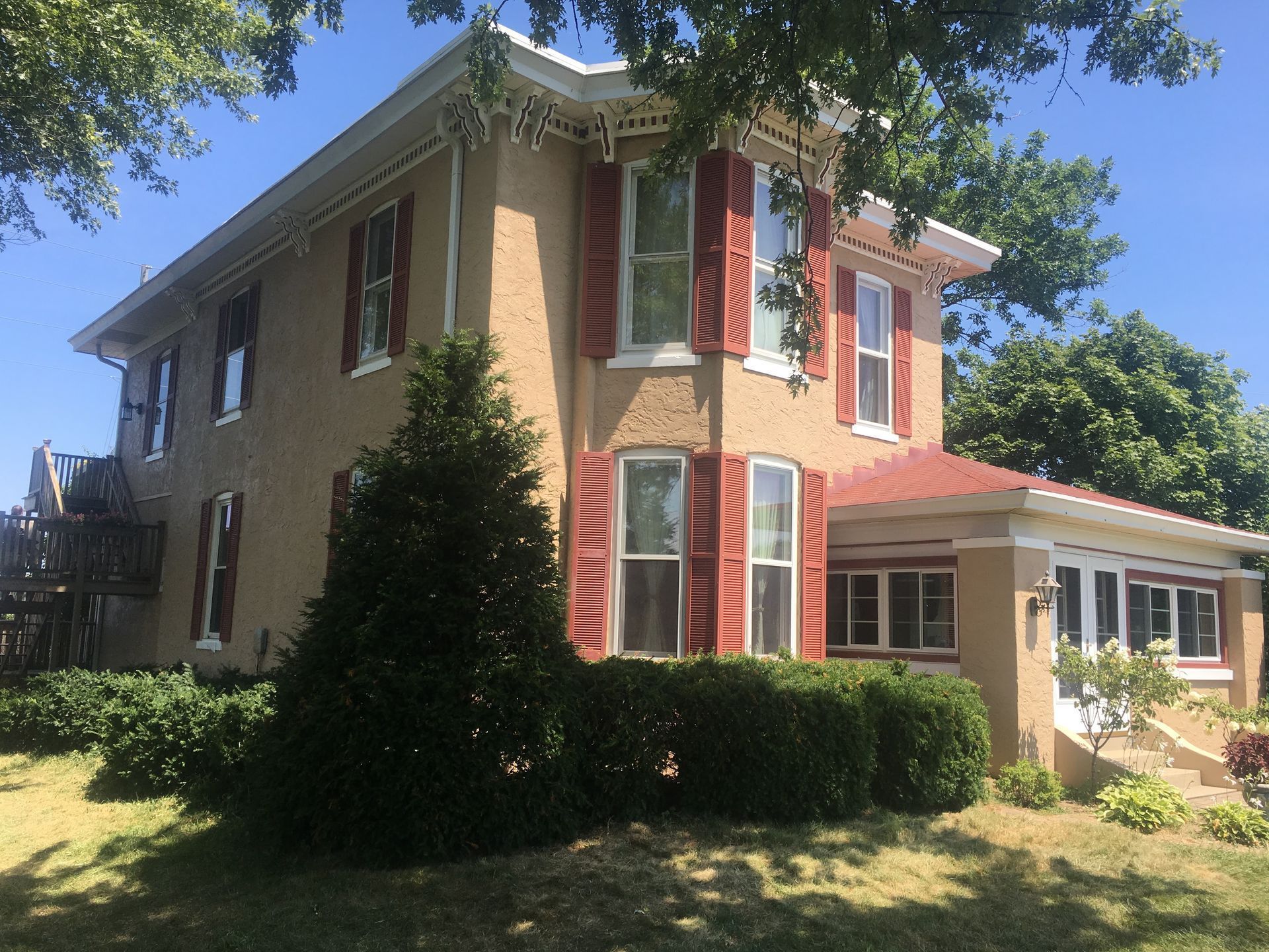 Two-story tan house with red shutters, white trim, and a red-roofed porch in a sunny yard