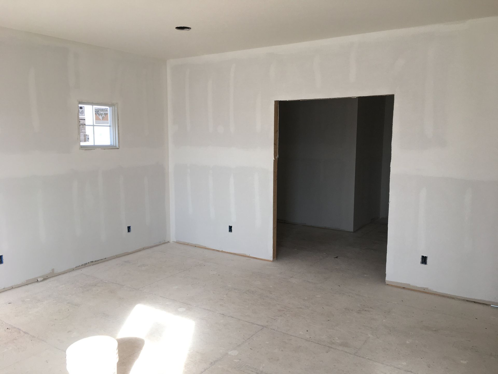 Empty drywall room with unfinished floor, doorway, and small window letting in sunlight