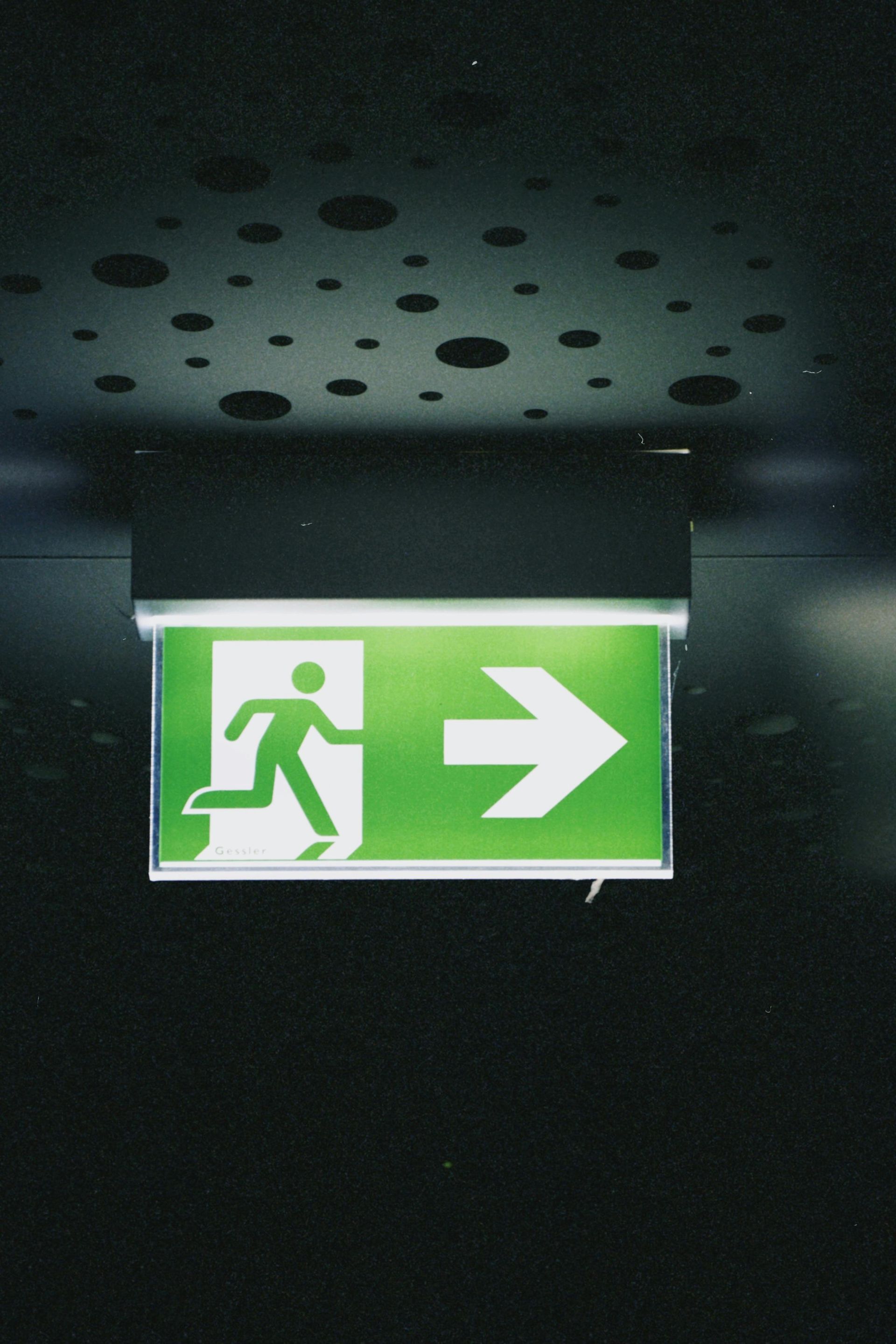 Illuminated green emergency exit sign with running figure and right-pointing arrow in a dark ceiling corridor