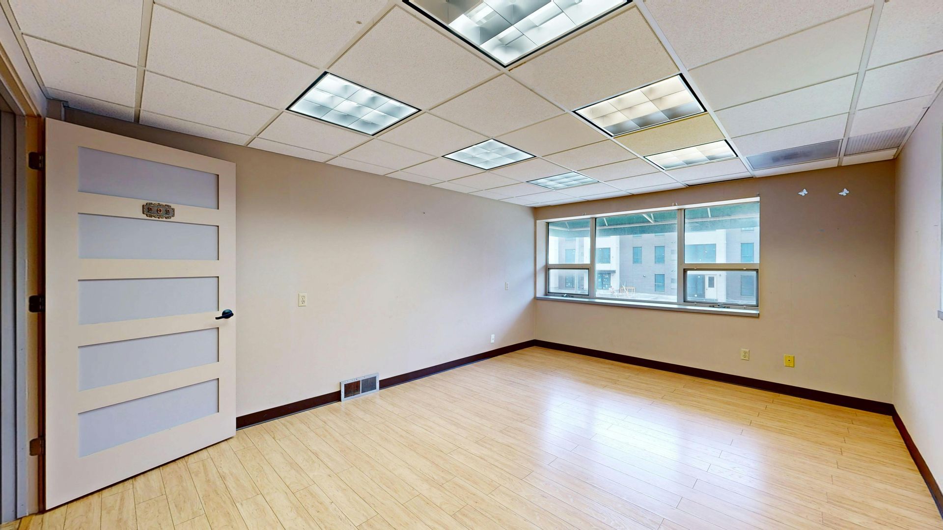 Empty office room with wood floors, beige walls, and large windows under fluorescent ceiling lights