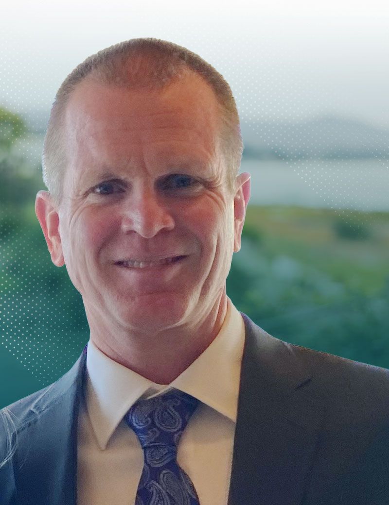 Smiling man in a suit and tie standing indoors with a blurred outdoor view behind him
