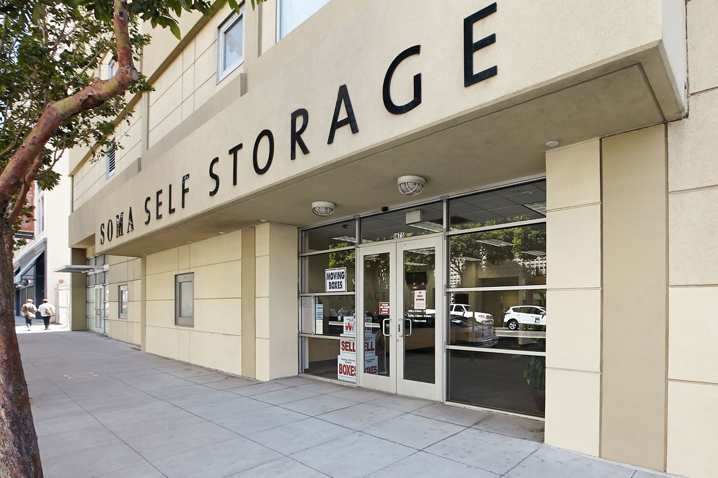 Exterior of a self-storage building with black lettering. Glass double doors lead inside. A tree is on the left.